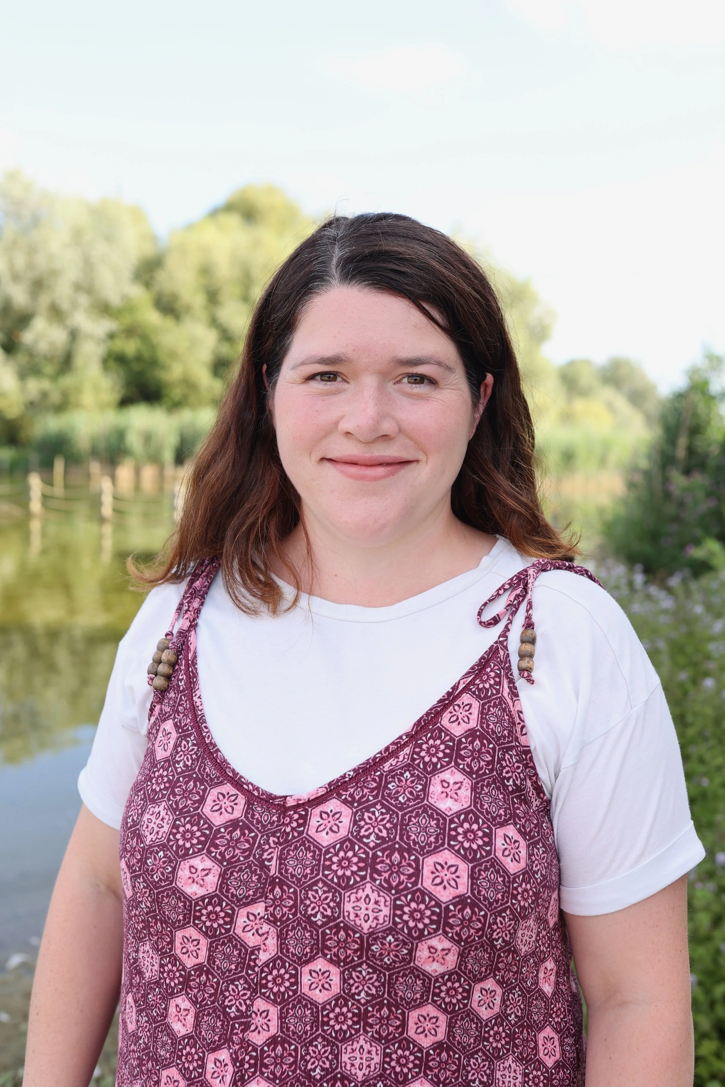 A woman with dark brown hair standing outdoors near a body of water, wearing a white t-shirt and a patterned pink and purple dress, with trees in the background.