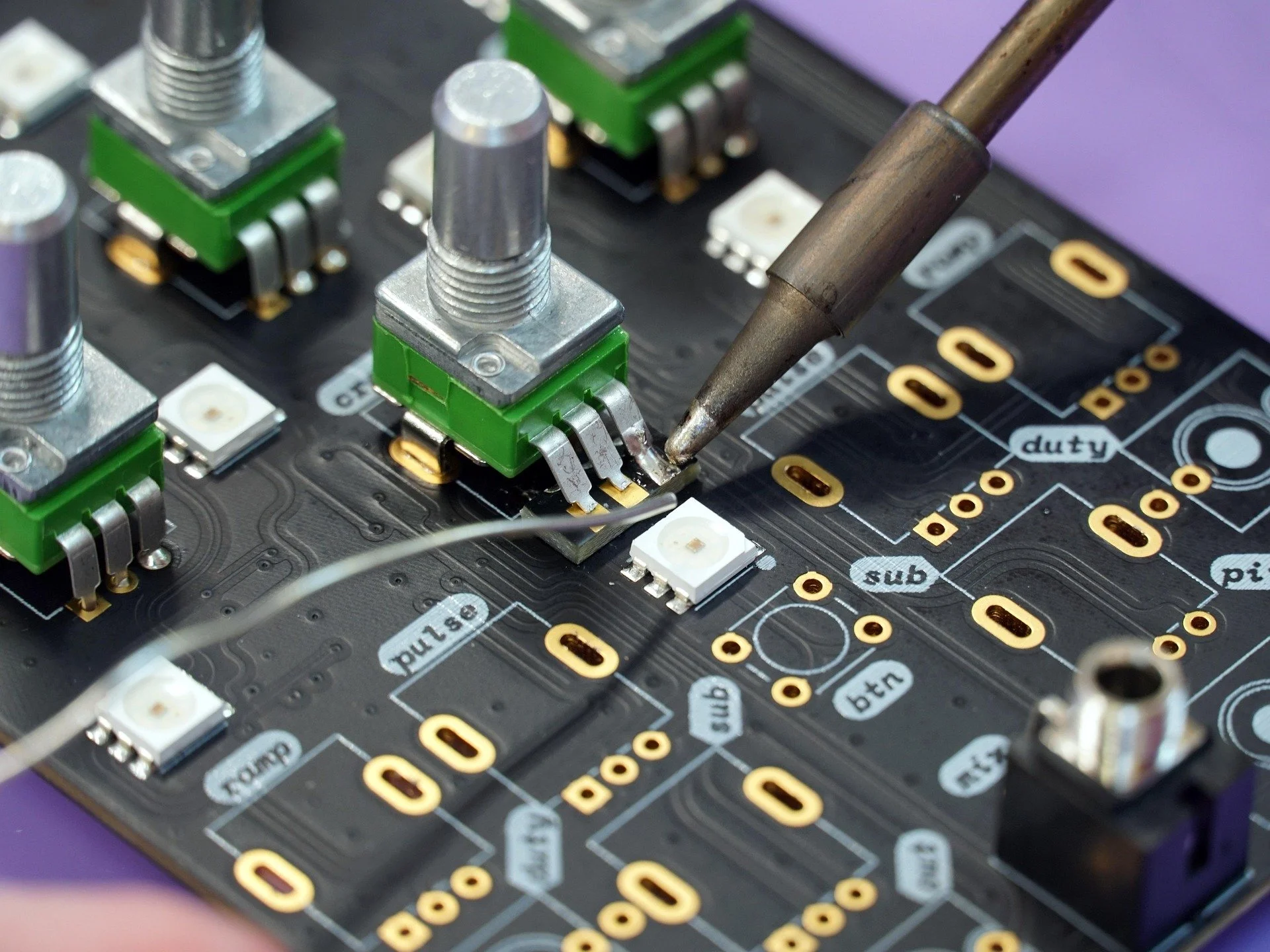 Close-up of a printed circuit board with electronic components, soldering iron, and labeled traces.