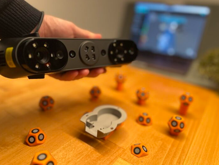 Close-up of a hand holding a black electronic device above a wooden table scattered with small orange and black geometric objects and a white circular component, with a blurry computer screen in the background.
