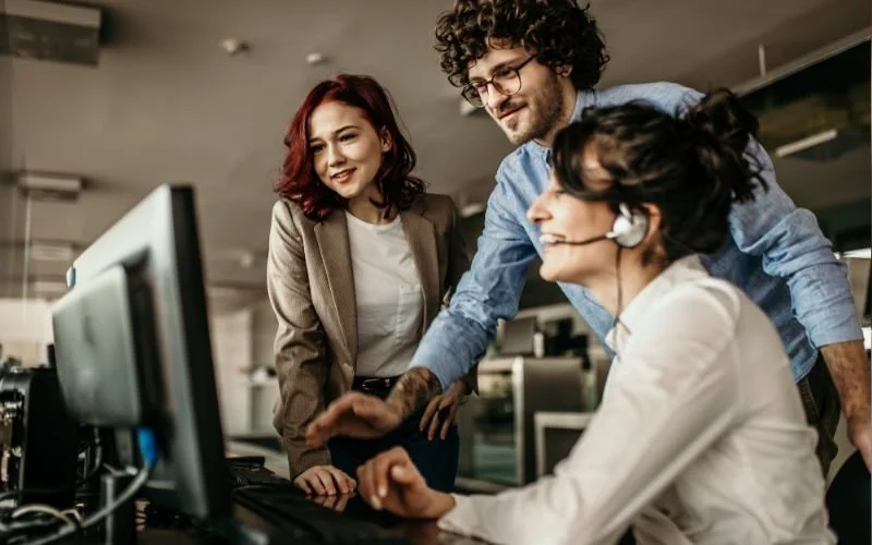 Three people working at a computer in an office, two standing and one sitting with a headset, all smiling and focused on the screen.