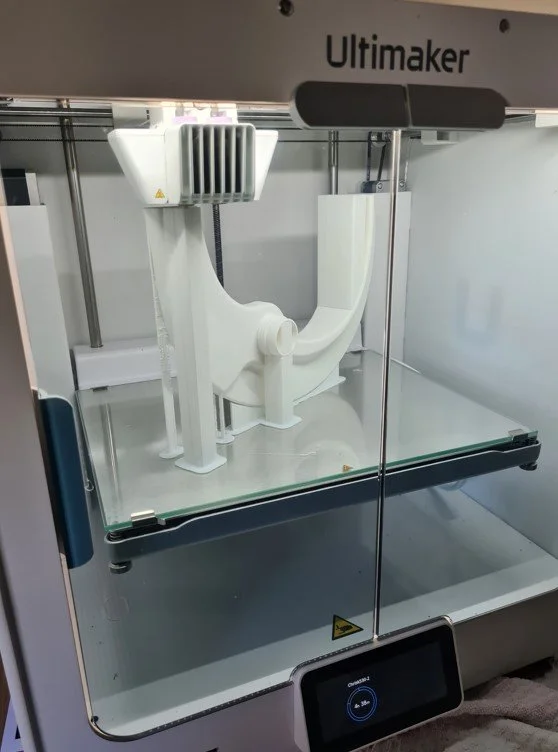 A white 3D printer labeled 'Ultimaker' printing a complex object in a clear enclosed case.