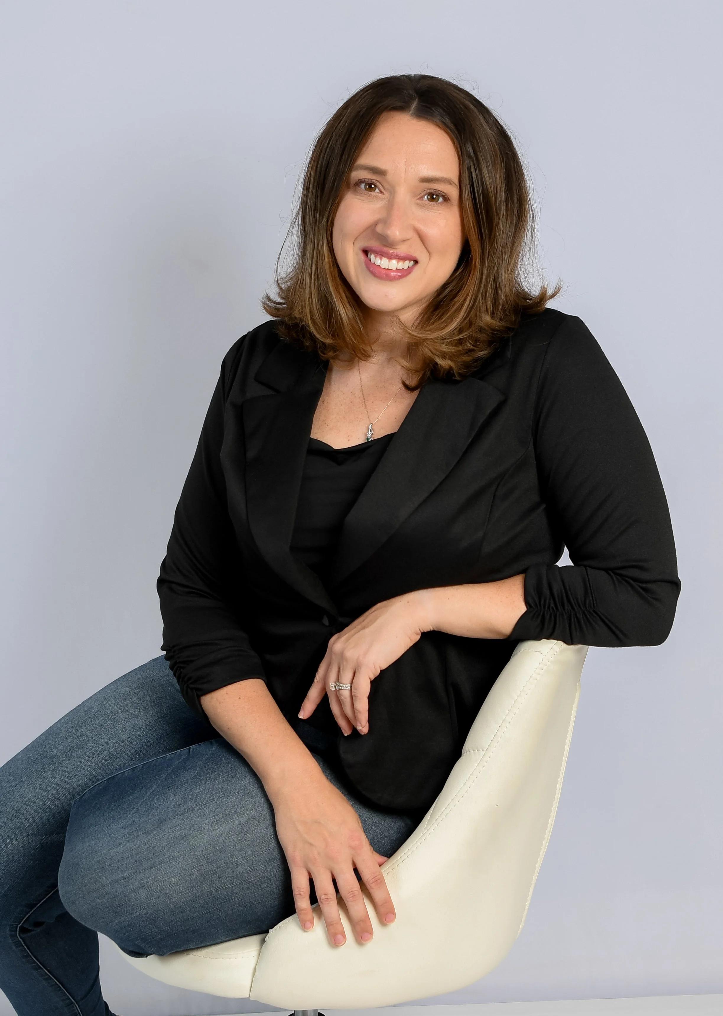 A woman with shoulder-length brown hair, smiling, wearing a black blazer and jeans, sitting on a white modern chair against a plain gray background.