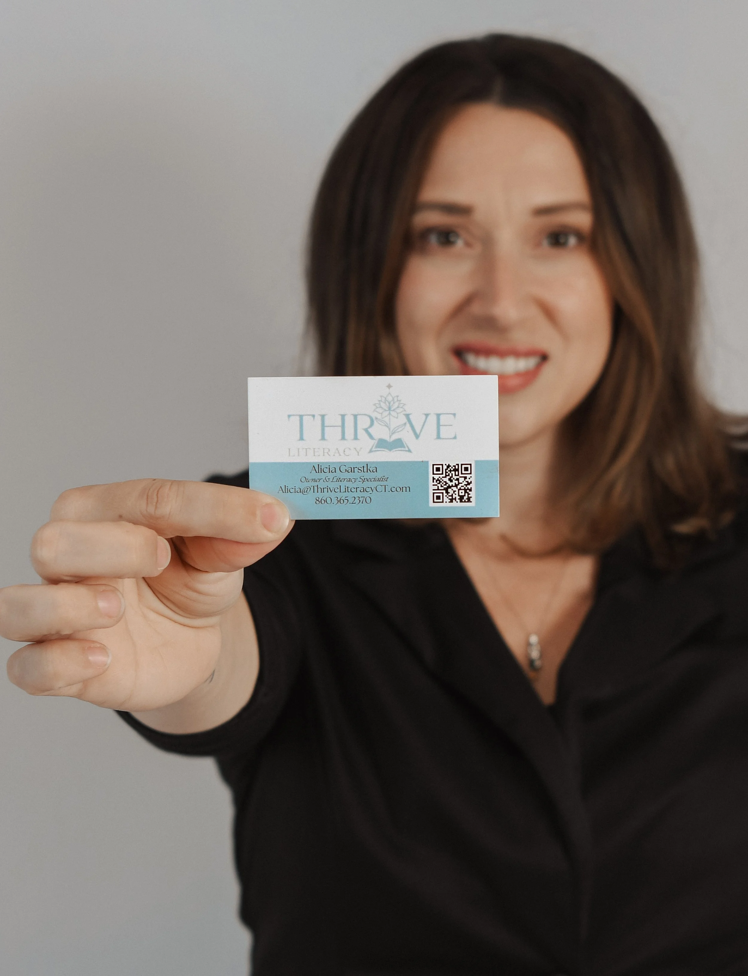 A woman with brown hair smiling and holding a business card towards the camera. The business card reads 'THRIVE Literacy', with contact details including a name, email, and phone number, as well as a QR code.