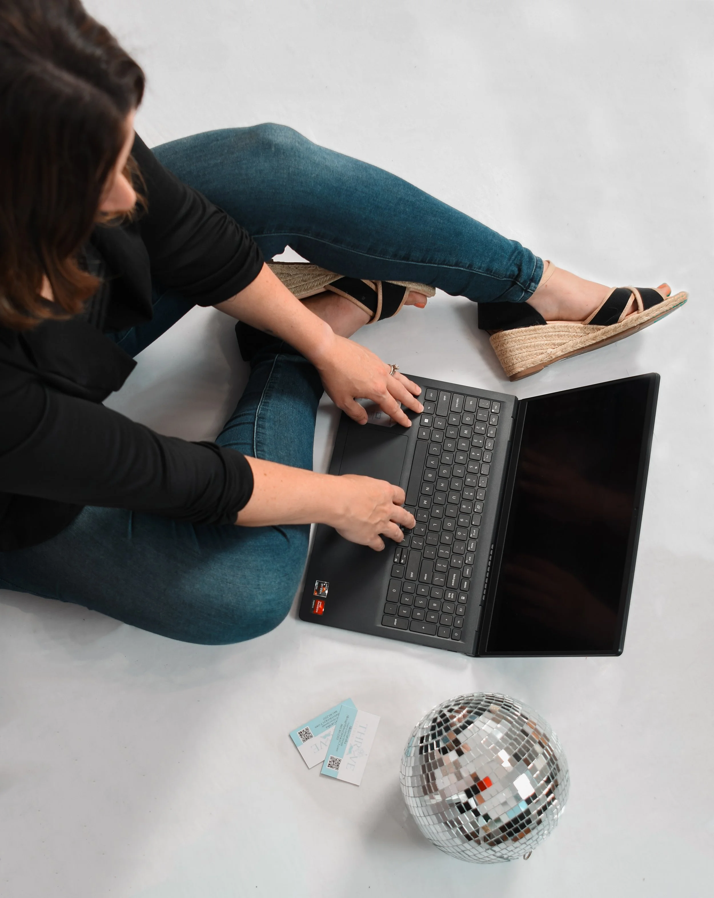 A woman sitting on the floor using a laptop, with a disco ball and tickets nearby.