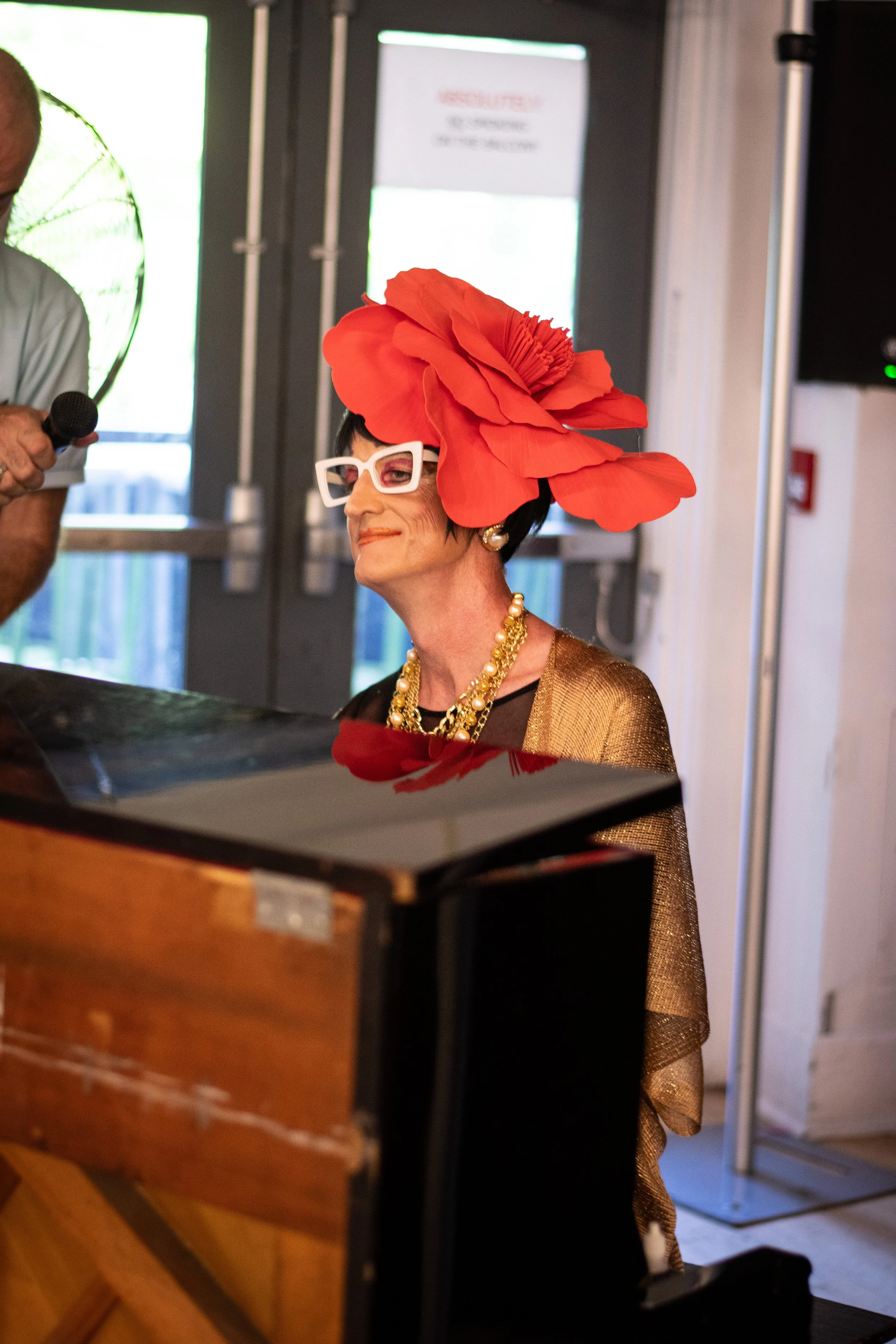 A Drag queen with a large red flower in her hair sits at a black upright studio piano facing her audience for a community building interactive performance.