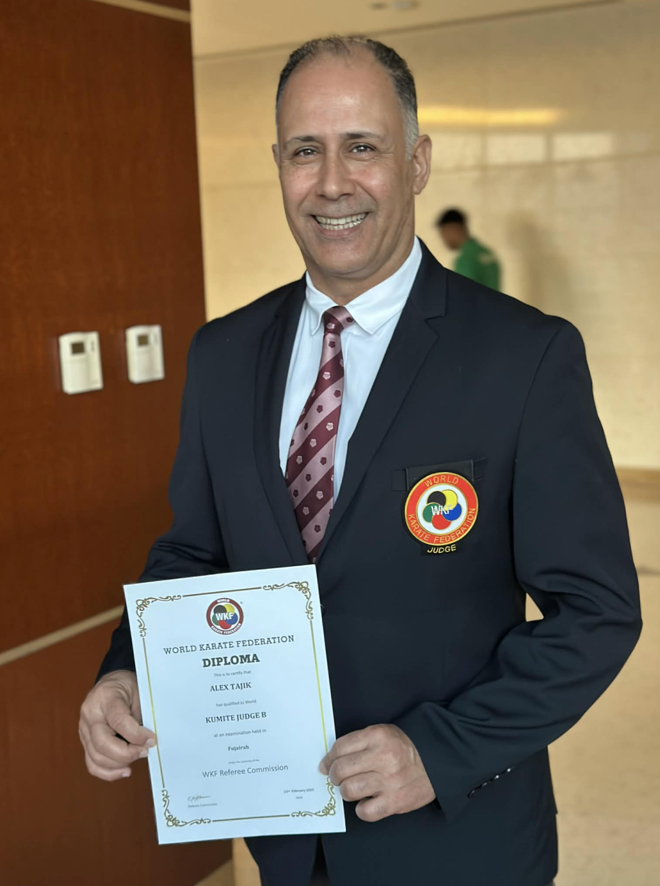 Sensei Alex Tajik from Tajik Karate Academy, a World Karate Federation–certified instructor, is pictured wearing a blazer with an official WKF patch, paired with a white dress shirt and maroon patterned tie.