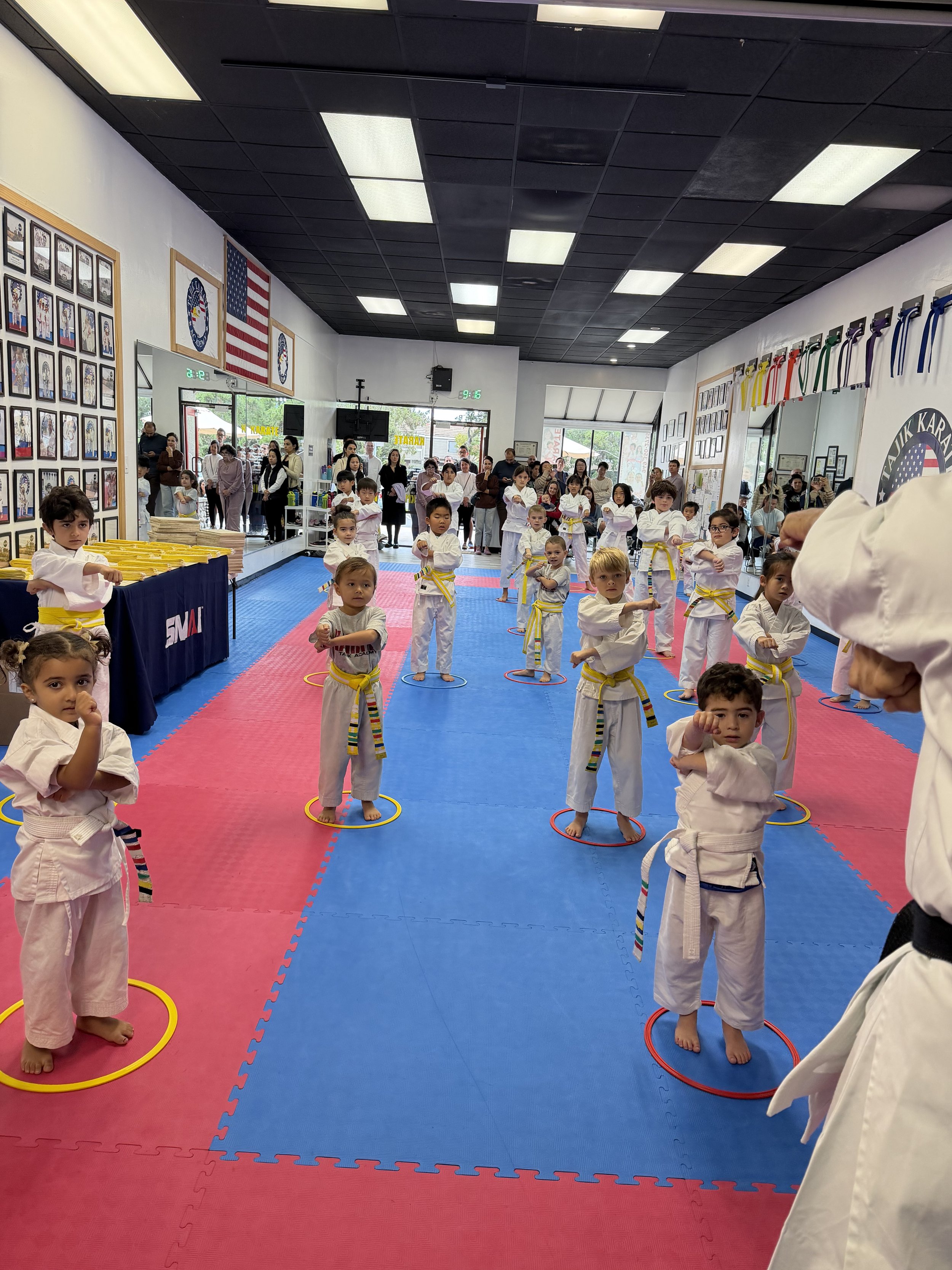 Children in martial arts uniforms practicing at a dojo, with spectators watching from the back.