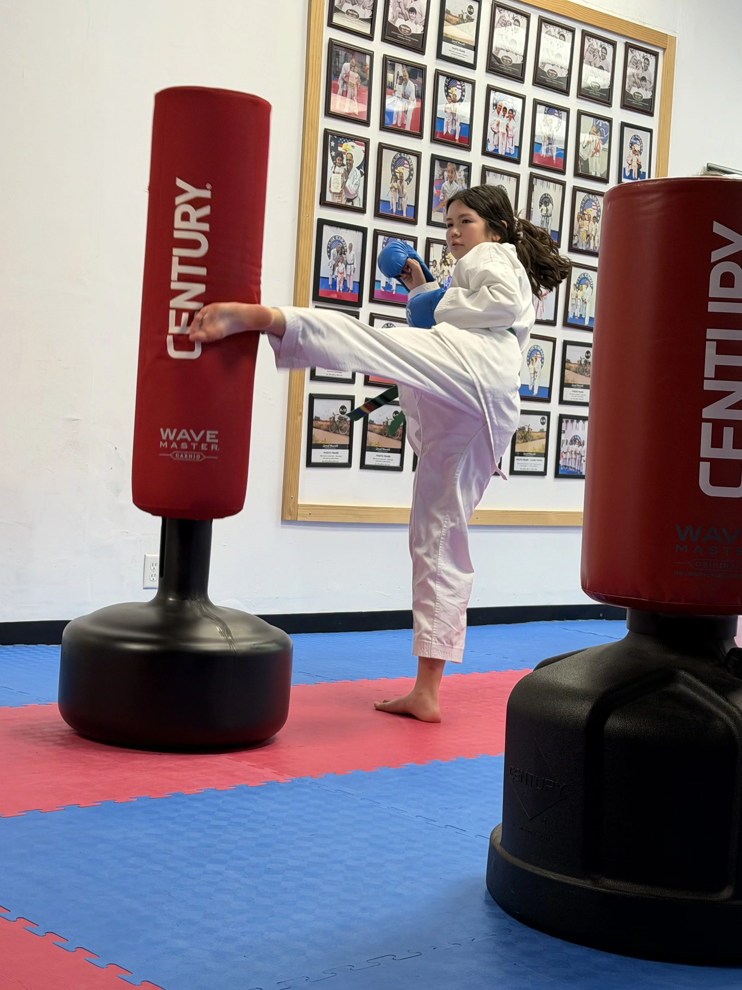 Young girl in a karate uniform practicing a high kick with a blue glove on her hand, kicking a red punching bag in a martial arts dojo with framed photos on the wall.