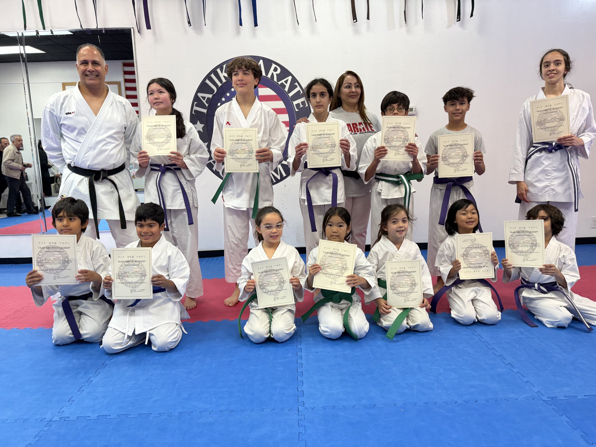 Group of children and two adults posing for photo in martial arts dojo. The children are wearing karate uniforms and holding certificates, some with colored belts indicating ranks. The adults are also in martial arts uniforms, one with a black belt. The background has a mirrored wall, a logo, and some martial arts banners.