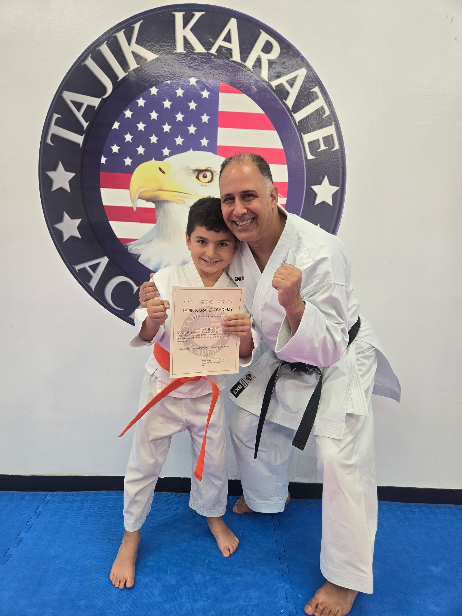 A young boy in a karate uniform with an orange belt, holding a certificate, standing next to a smiling instructor in a white karate gi with a black belt. They are posing in front of a large Tae Kwon Do Academy crest featuring an eagle and the American flag.