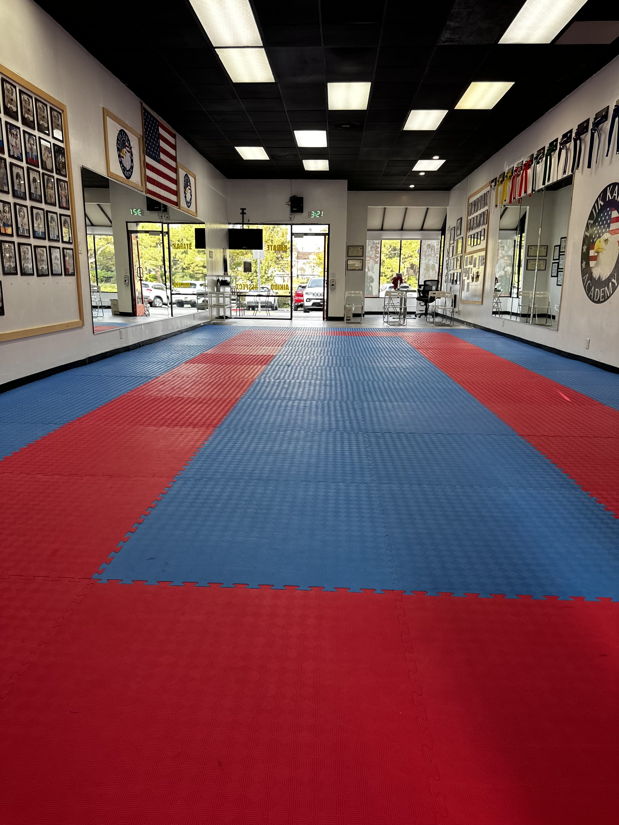 Interior view of a martial arts dojo with red and blue mats, framed photos on the wall, American flags, and a front entrance with large windows and glass doors.