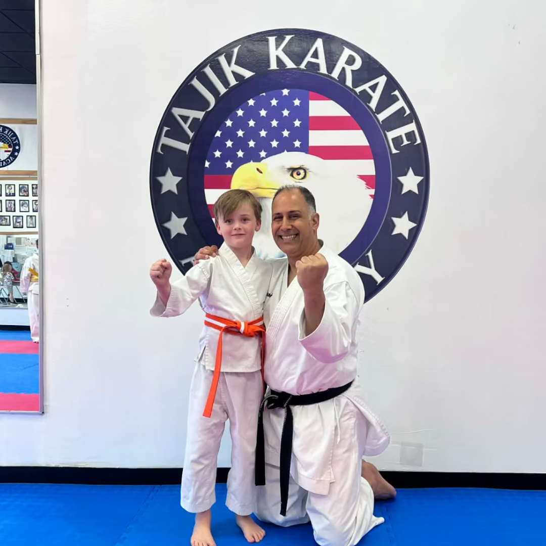 A young boy and an adult man in martial arts uniforms pose together inside a martial arts dojo, smiling and making fists. Behind them is a large sign with a bald eagle head and the American flag, with the words 'TAJIK KARATE'.