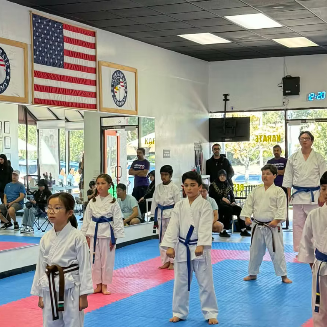 Children practicing karate in a dojo, wearing white gi uniforms with colored belts, standing on red and blue mats. The dojo has American flags and martial arts banners on the wall, with spectators watching from the background.