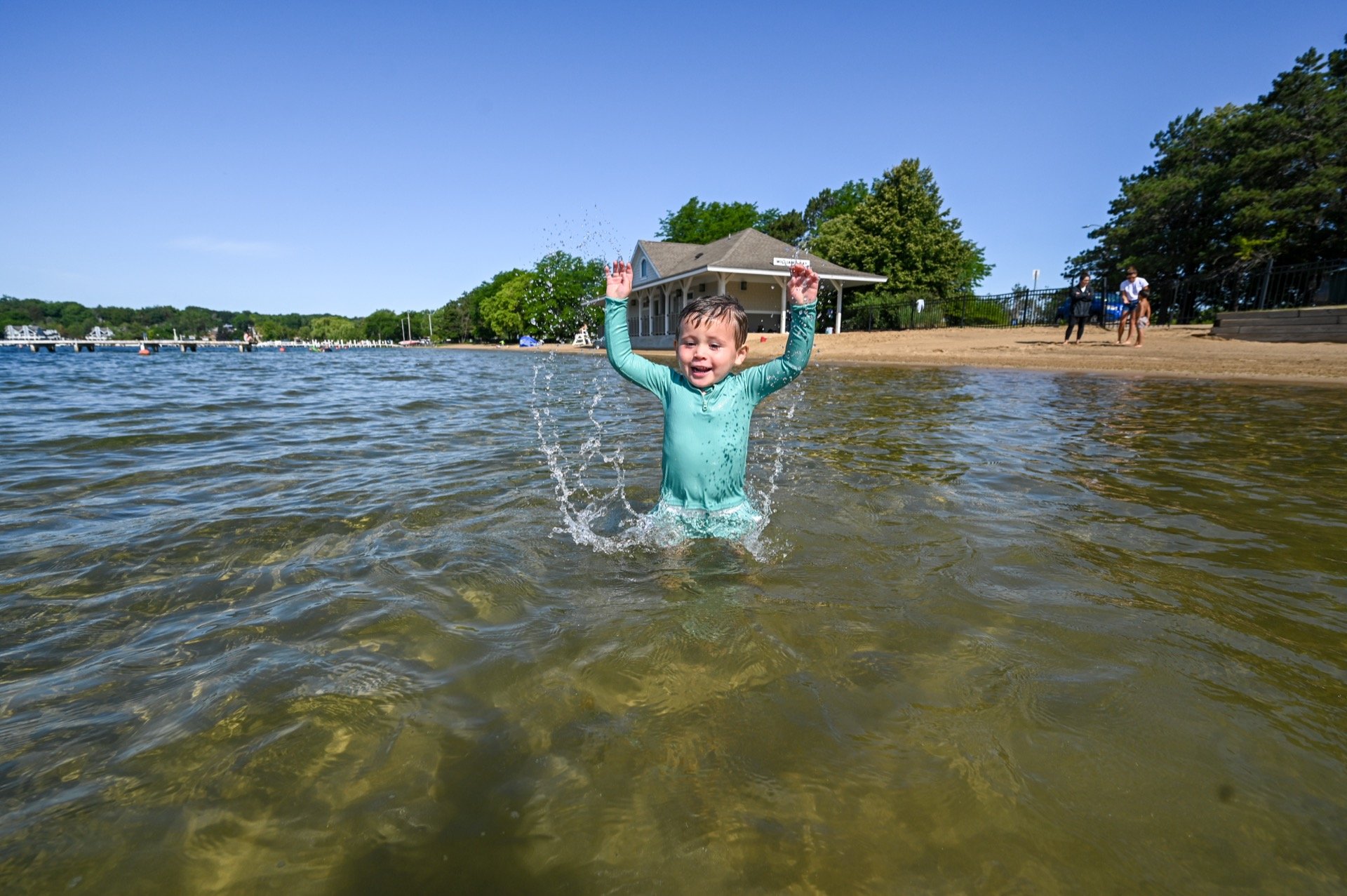 A young boy in a turquoise rash guard enjoys playing in shallow water at a lakeside beach, smiling as water splashes around him on a sunny day.