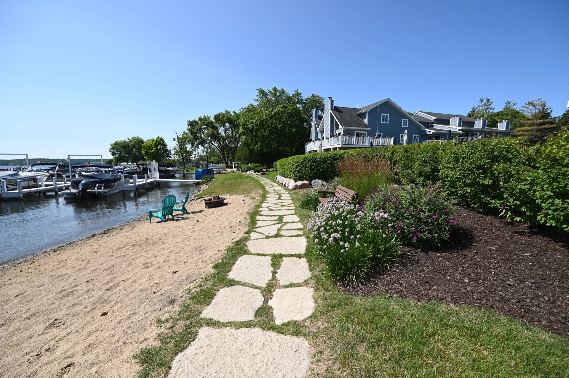 A sunny lakeside scene with a stone pathway, sandy beach, green bushes, colorful flowers, and houses with balconies in the background.