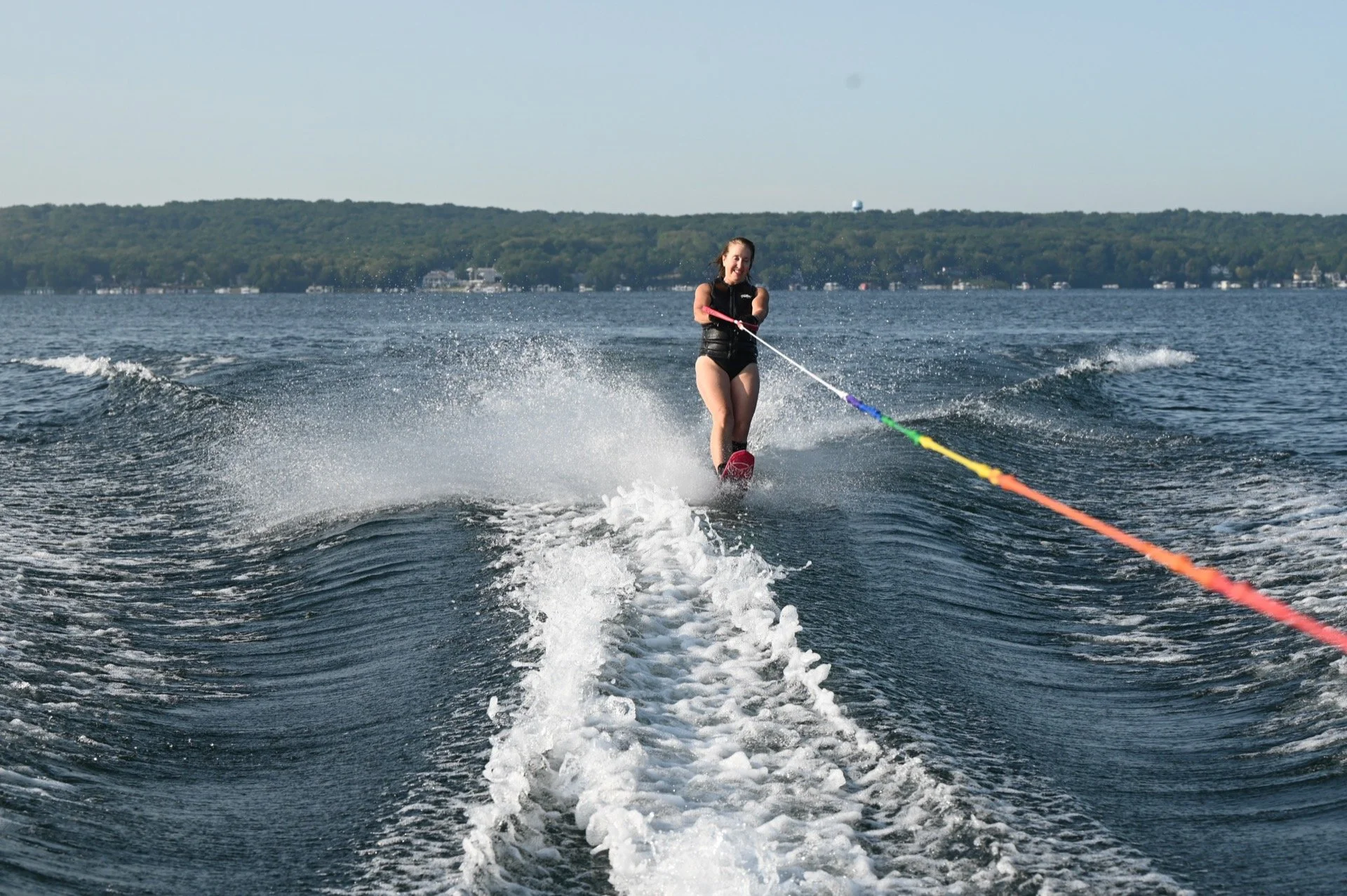 A woman in a black swimsuit wakeboarding on a lake with a forested shoreline in the background, smiling, holding a handle connected to a colorful tow rope, creating wake and spray.