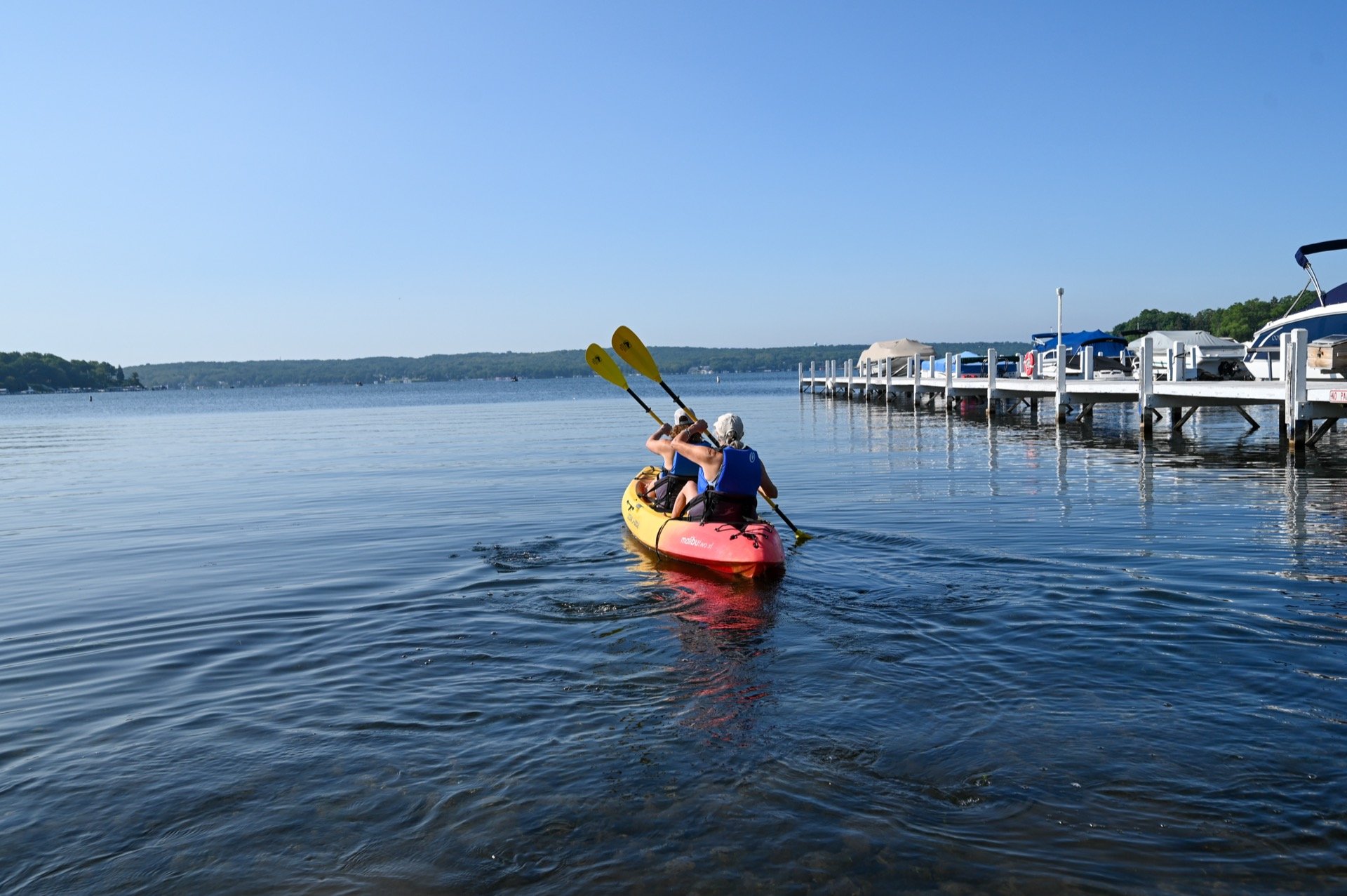 Two people kayaking on a calm lake near a dock with boats and jet skis, under a clear blue sky.