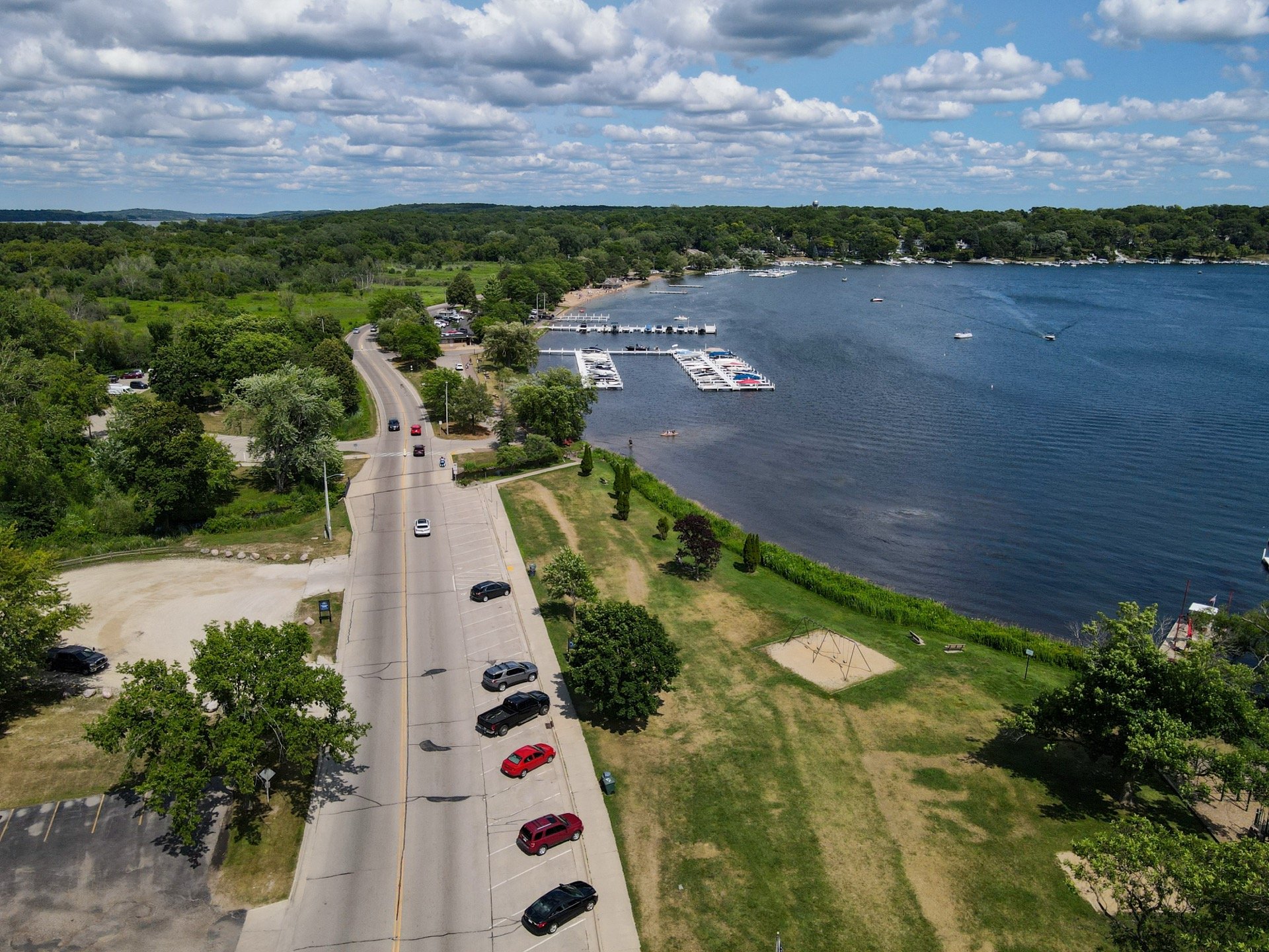 An aerial view of a lakeside area showing a parking lot, a road with cars, a grassy park with trees, a playground, and a marina with boats docked in the water. The lake is surrounded by green trees and hills under a partly cloudy sky.
