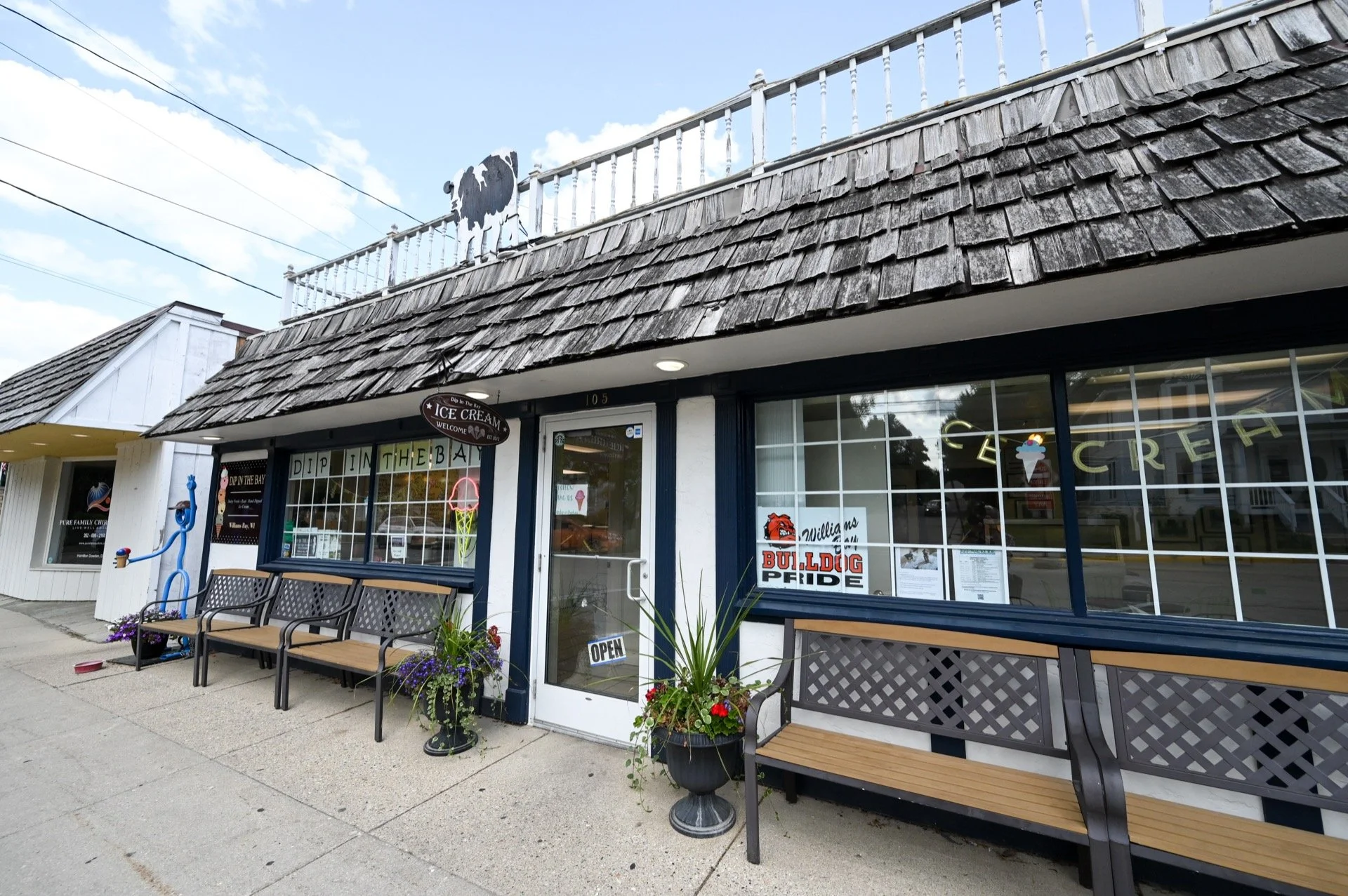 A small ice cream shop on a sidewalk with benches and potted plants outside. The shop has large windows with signs and a door marked 'Open,' and a sign above the entrance advertising ice cream. The building has a shingled roof and a white railing on top.
