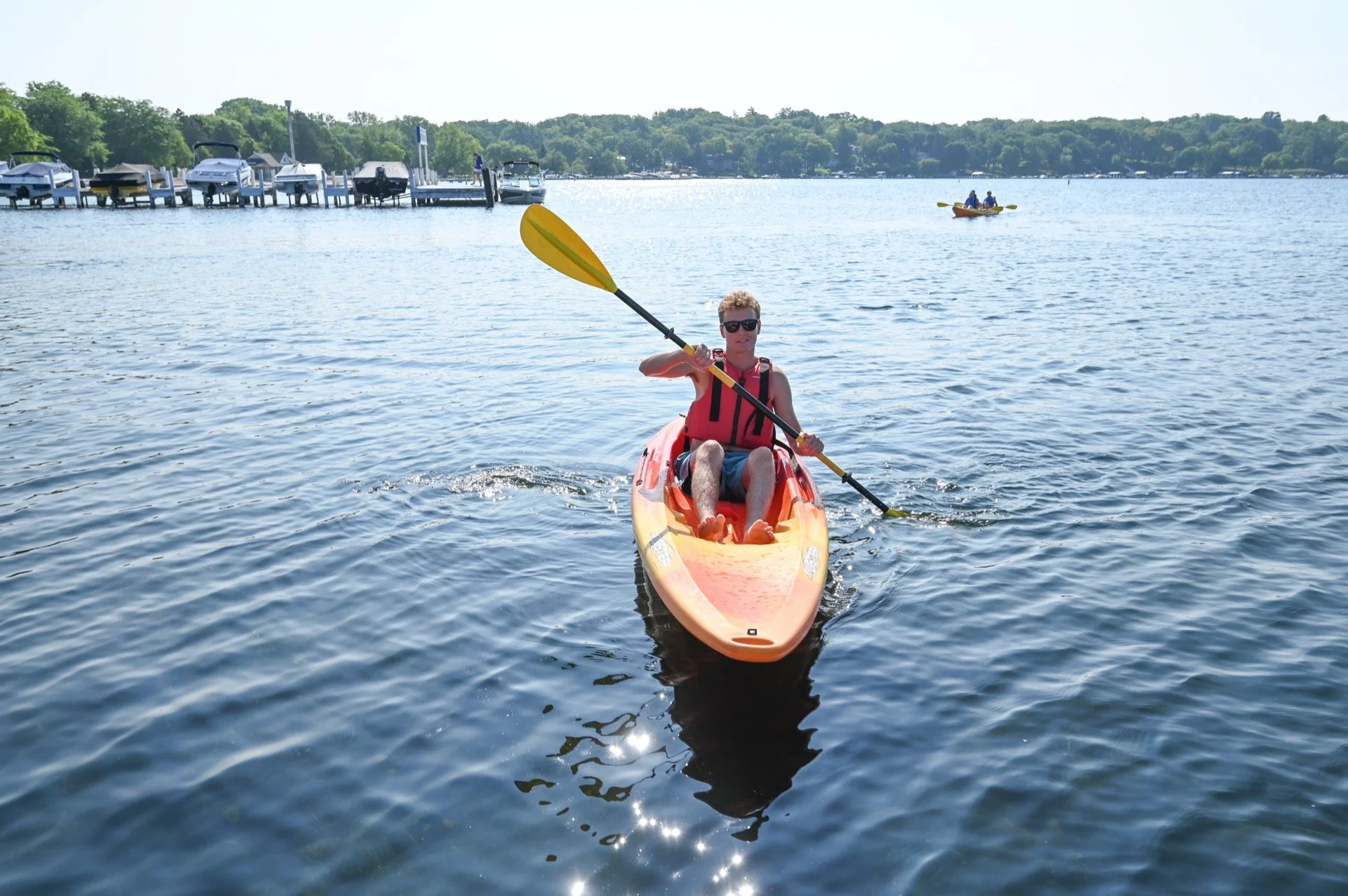 A young man kayaking on a lake, wearing sunglasses and a red life jacket, with boats and a dock in the background.