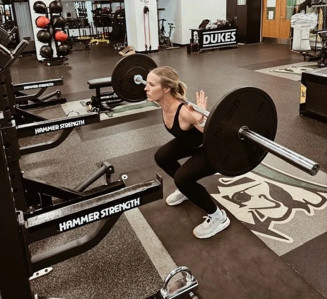 Woman performing a barbell squat exercise in a gym, with weights and fitness equipment around her.