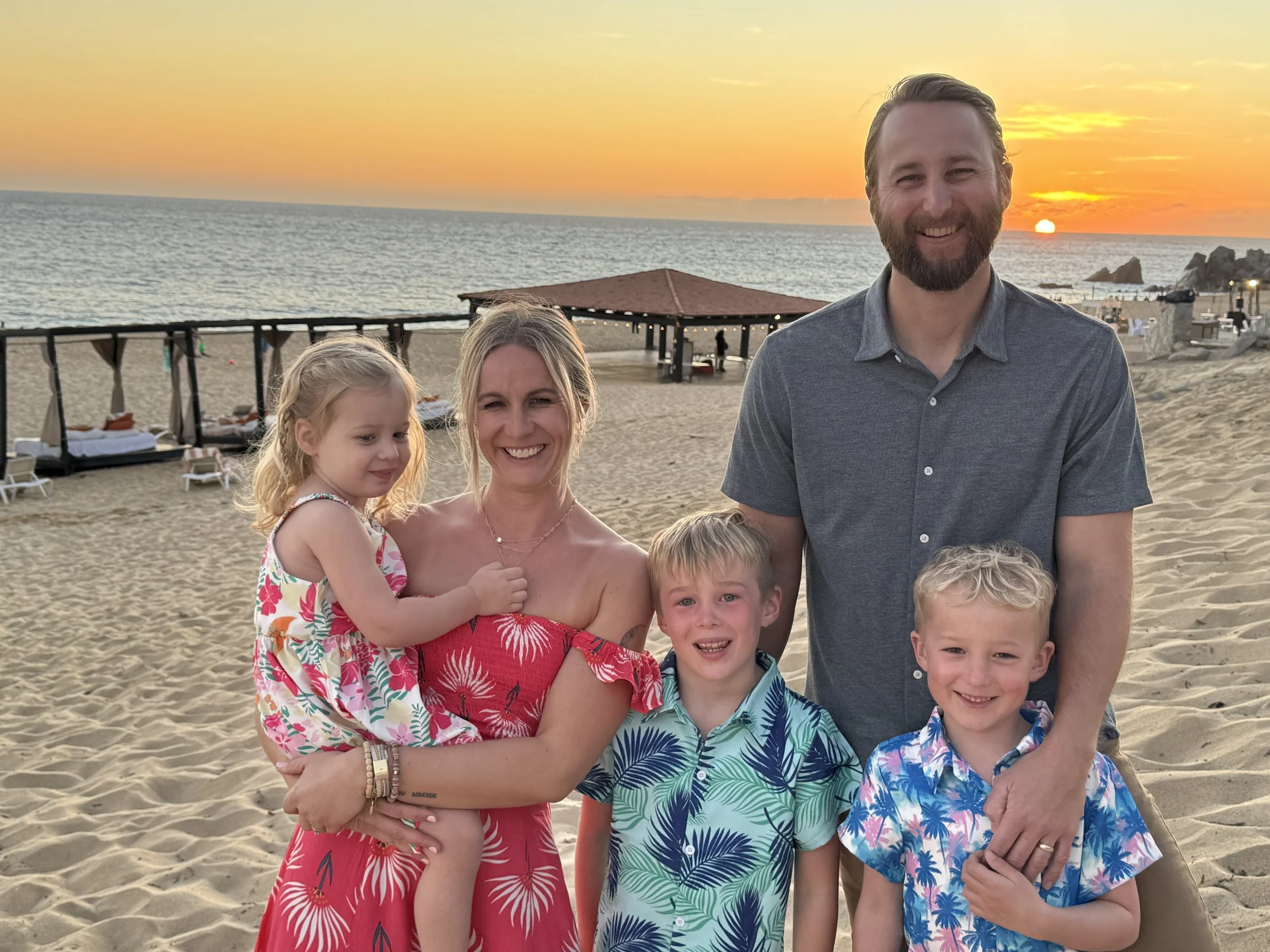 A family of five stands on a sandy beach at sunset. The mother is holding a young girl, and two boys stand in front. The father is standing to the right. The background includes a gazebo, lounge beds, and the ocean with the sun setting over the horizon.
