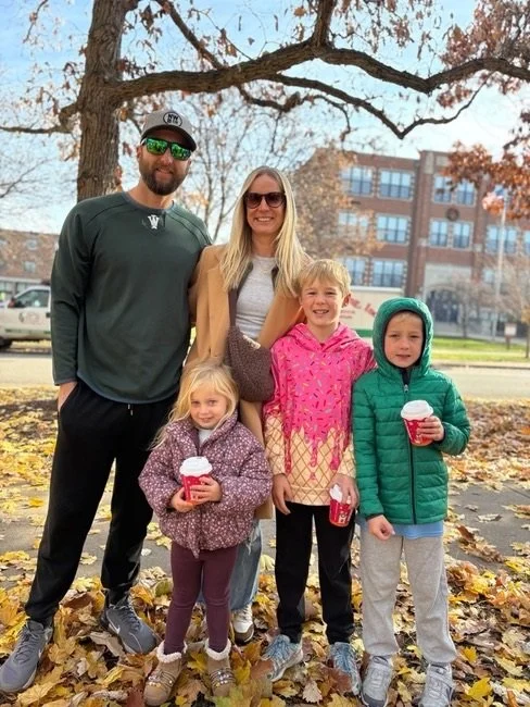 Family of five standing outdoors among fallen autumn leaves, smiling, with a large tree and a brick building in the background. The family has a man, woman, and three children, one girl and two boys, all dressed in casual fall attire, holding coffee cups.