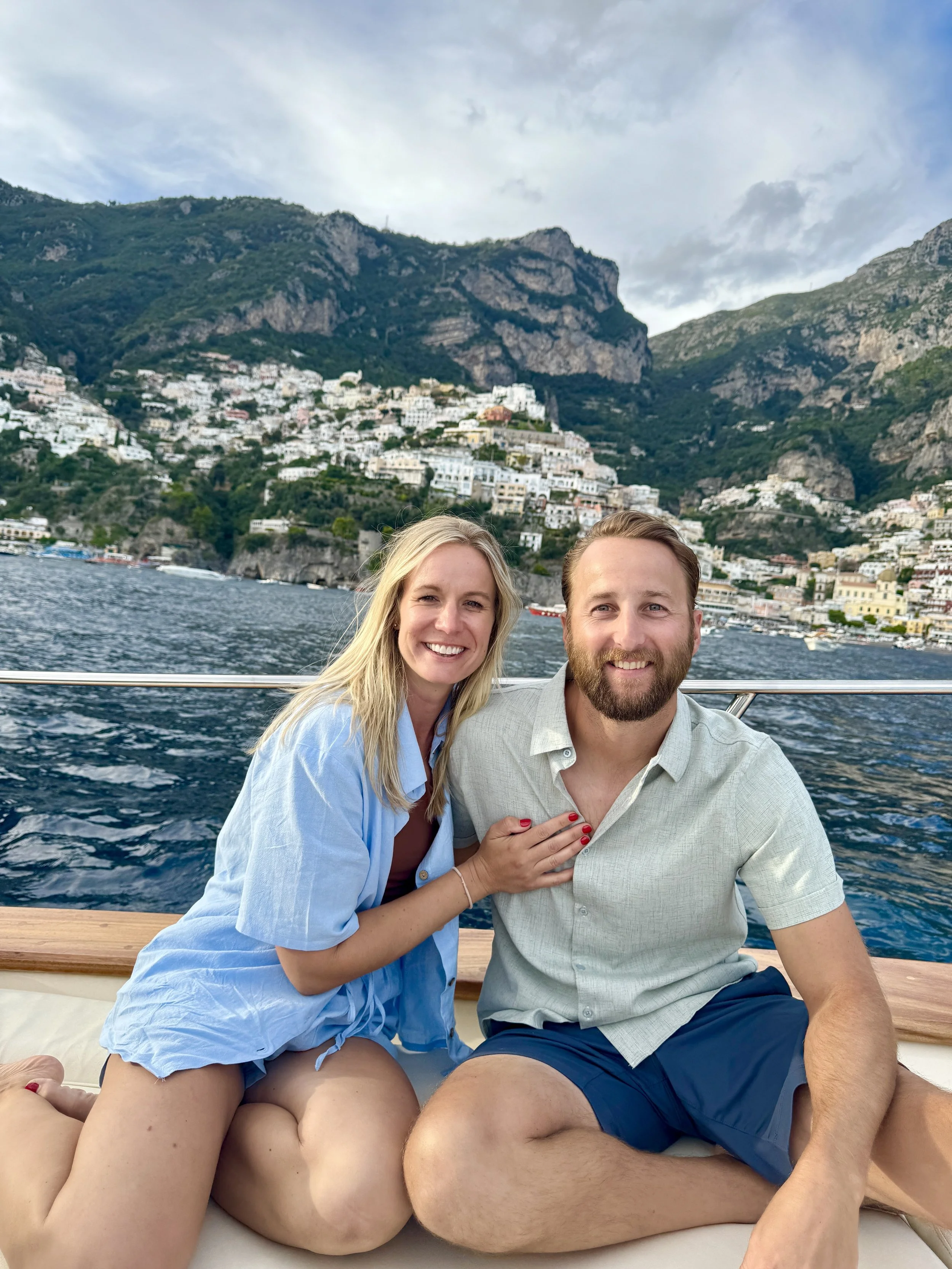 A smiling couple on a boat with a scenic seaside hillside town in the background.