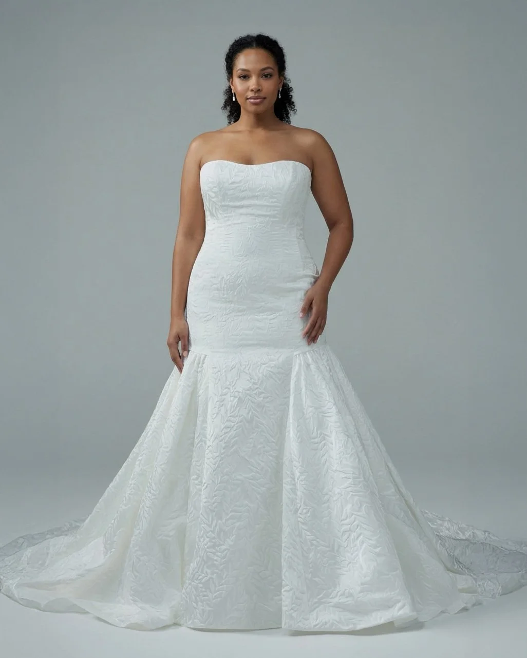 A woman wearing a strapless white wedding gown with a textured fabric and a flared bottom, standing against a plain gray background.