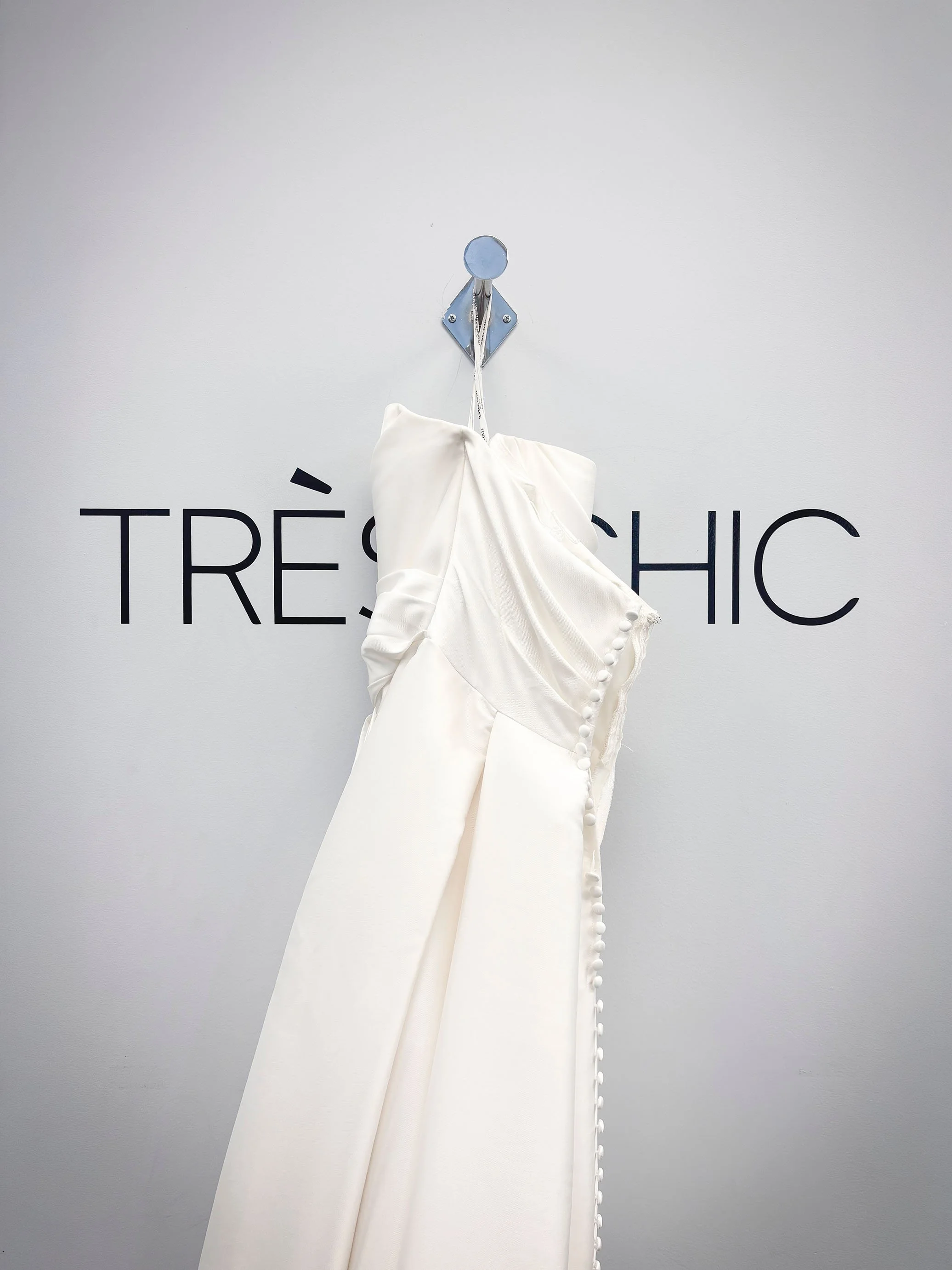A white wedding dress hanging on a wall hook with the brand name 'TRÈS CHIC' written on the wall behind it.