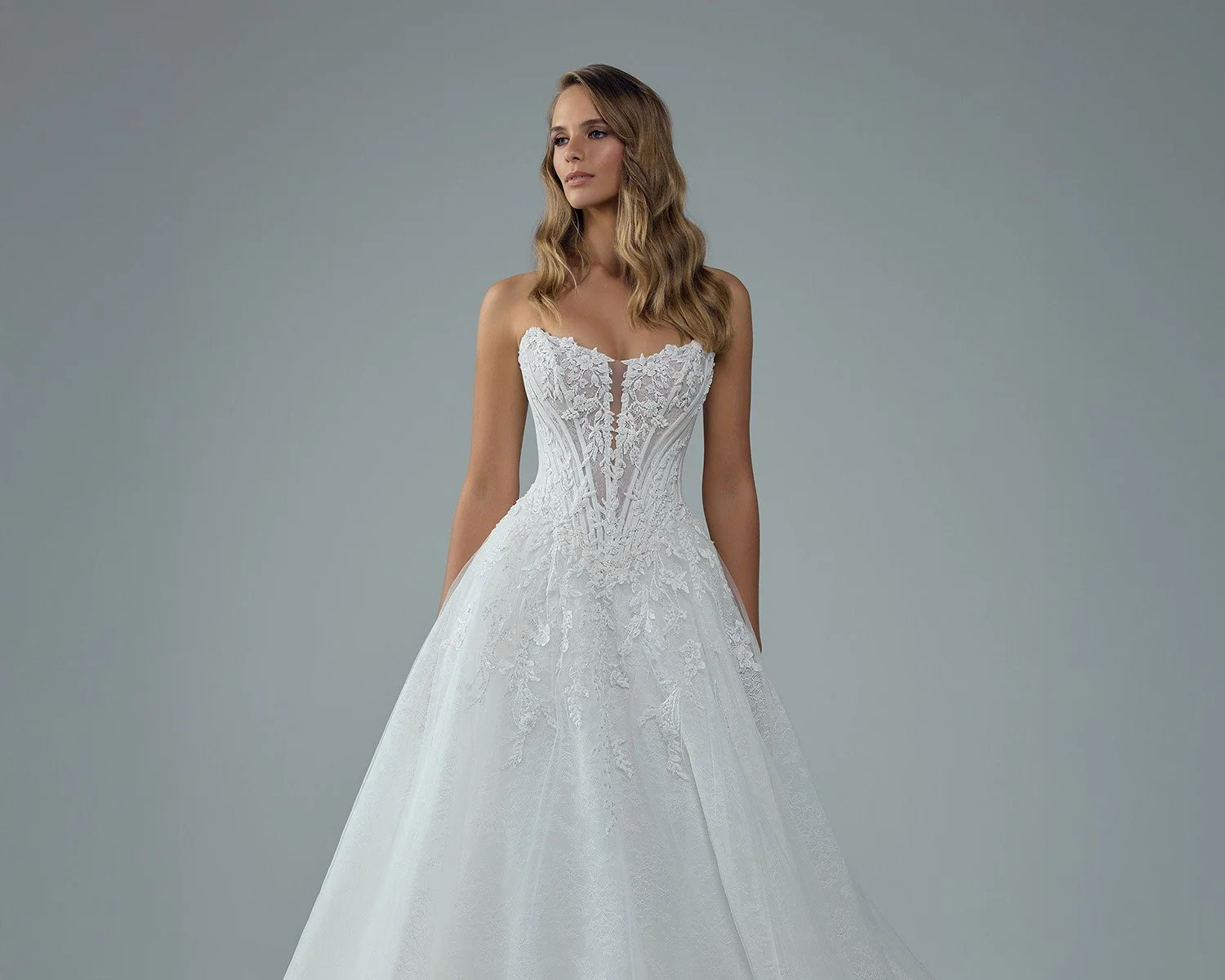 A woman in a strapless white wedding gown with lace detailing, standing against a gray background.