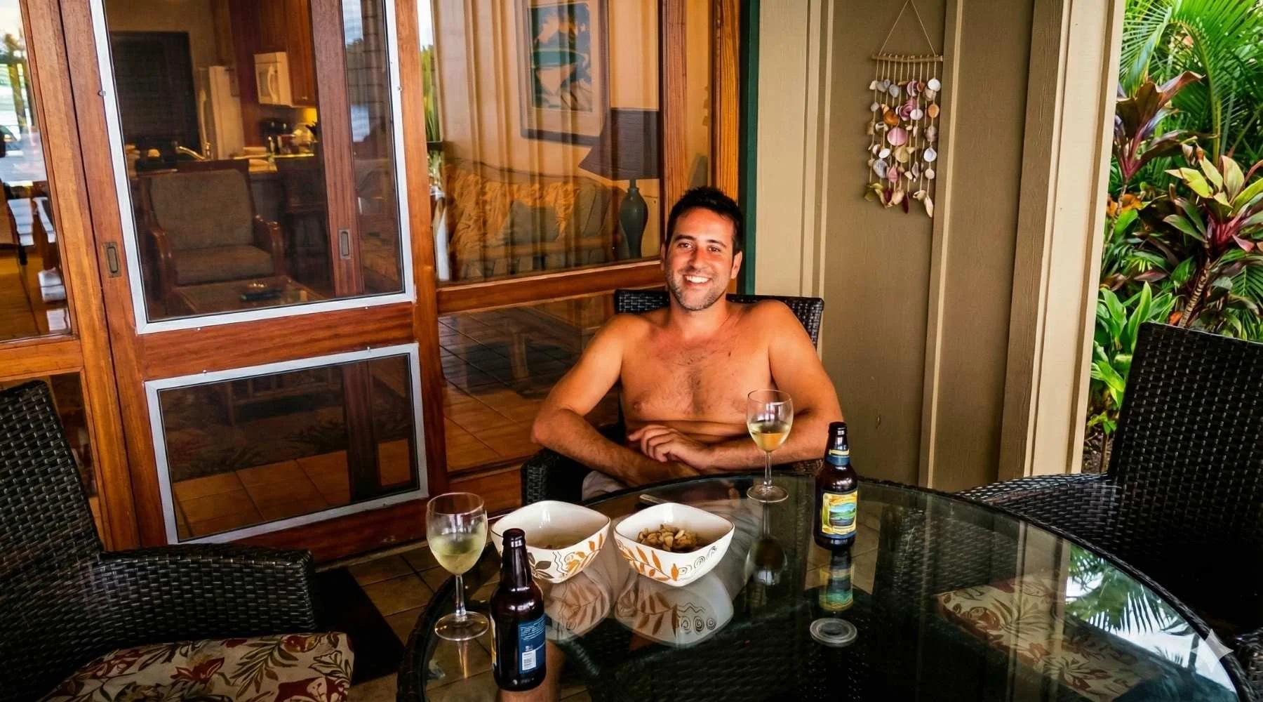 A shirtless man sitting at an outdoor wicker table with drinks and snacks, smiling.