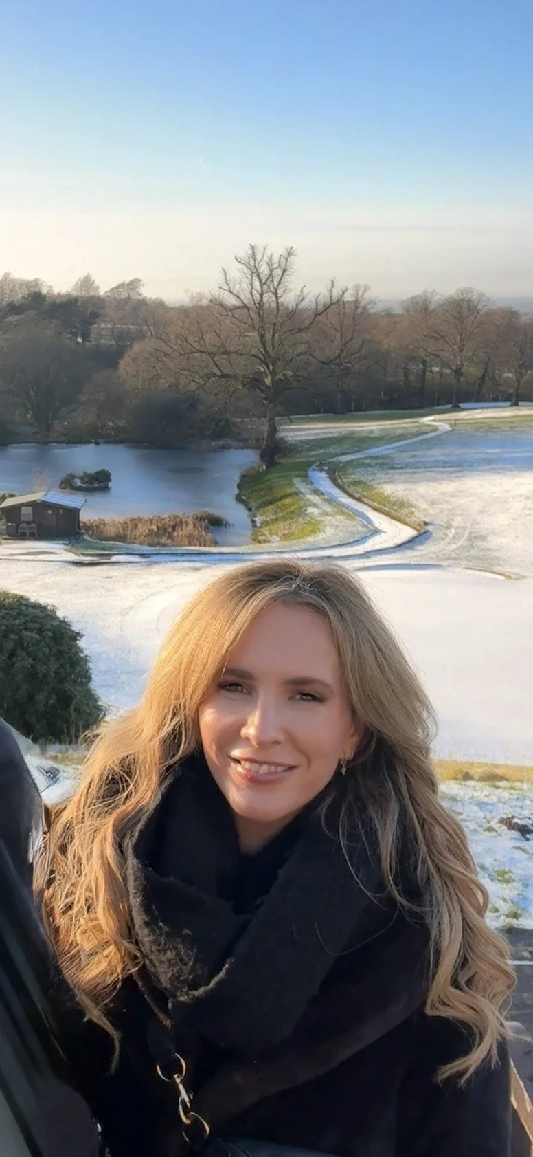 Young woman with long, wavy blonde hair smiling outdoors in winter with snow-covered ground, a small pond, and trees in the background.