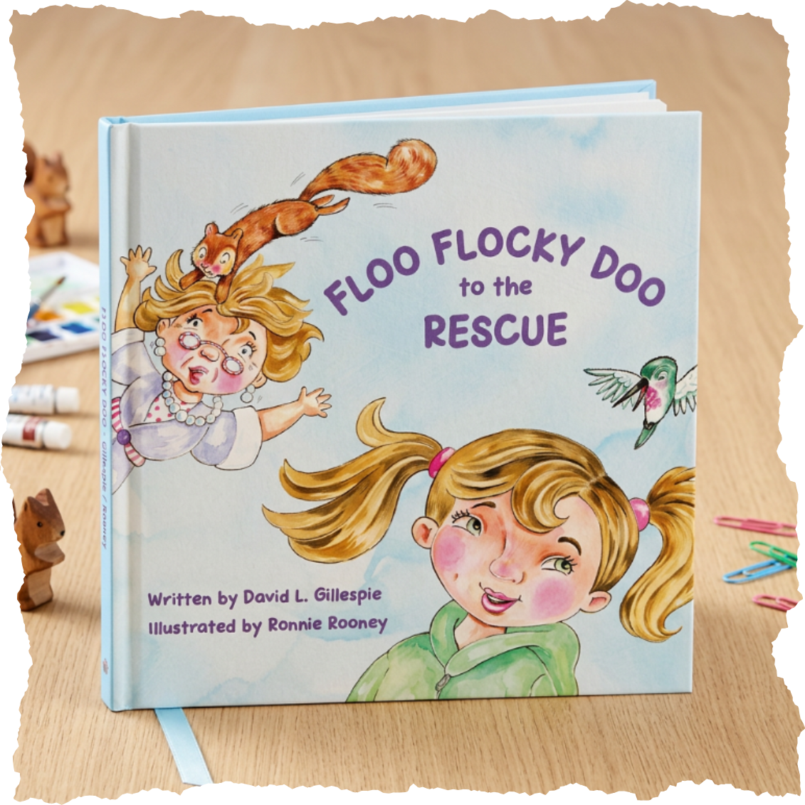 A children's book titled 'Floo Flocky Doo to the Rescue' with colorful illustrations of a girl with pigtails, a flying squirrel, a hummingbird, and a woman with glasses. The book is on a wooden table surrounded by art supplies.