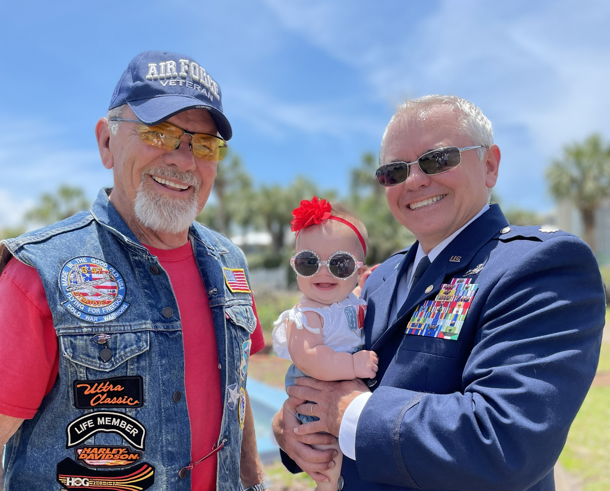 Two men in military uniforms holding a smiling baby girl with sunglasses and a red bow, outdoors on a sunny day.