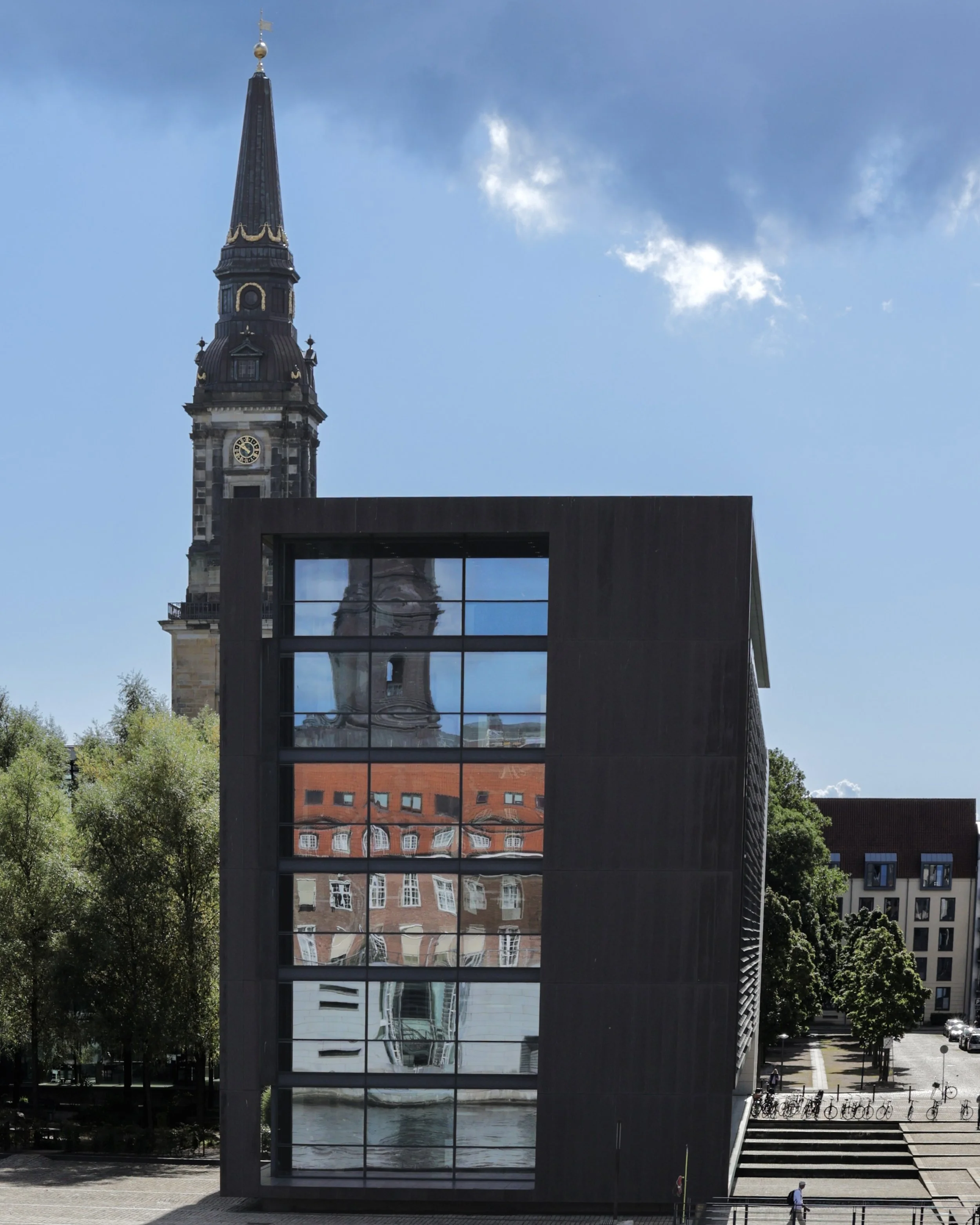 Modern black building with large glass windows reflecting a historic church tower with a clock and a red-tiled roof surrounding buildings, trees, and a partly cloudy sky.
