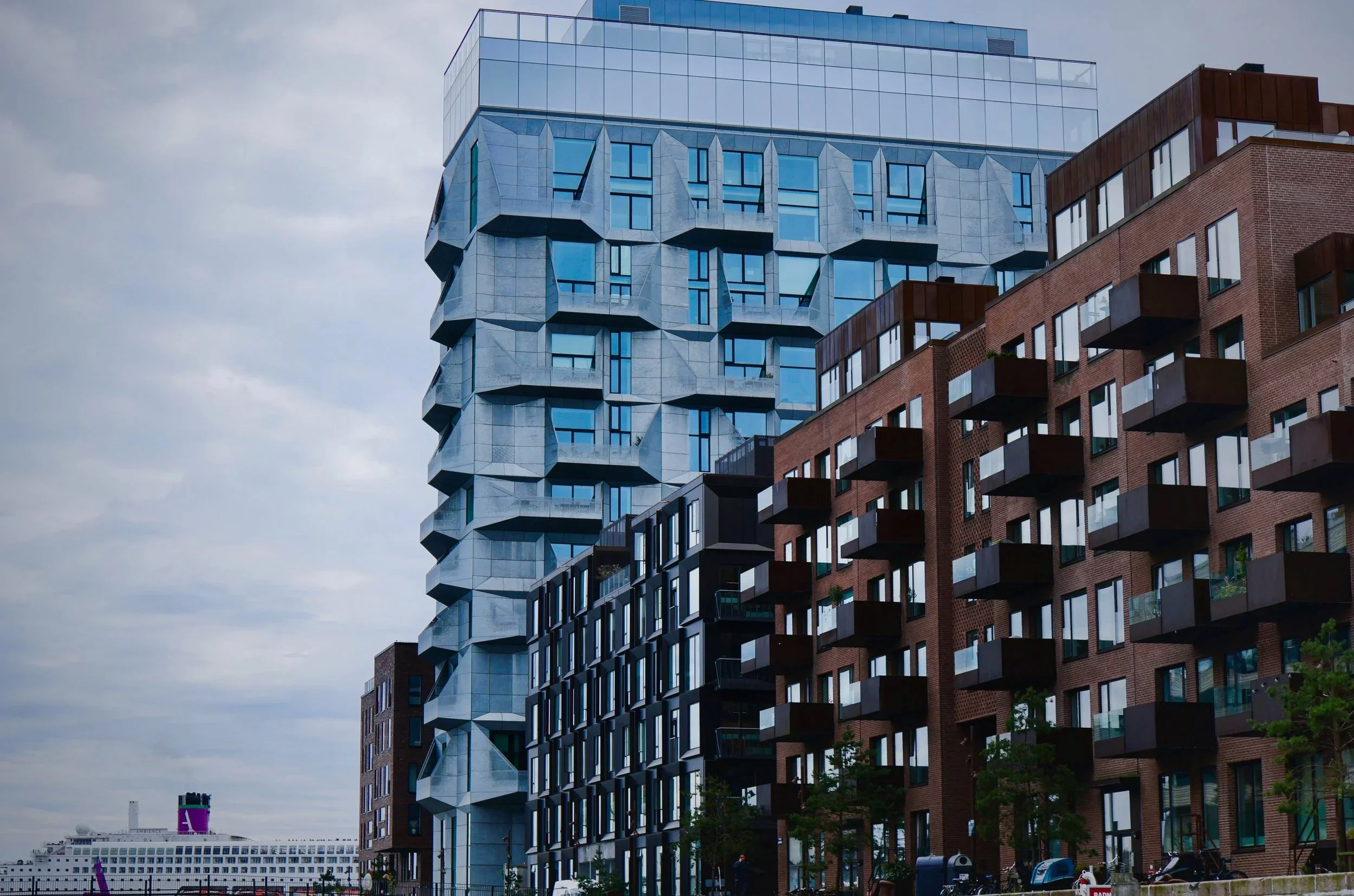 Modern high-rise buildings with a distinctive blue and gray geometric design on their facades, located in an urban setting with a ship visible in the distance.
