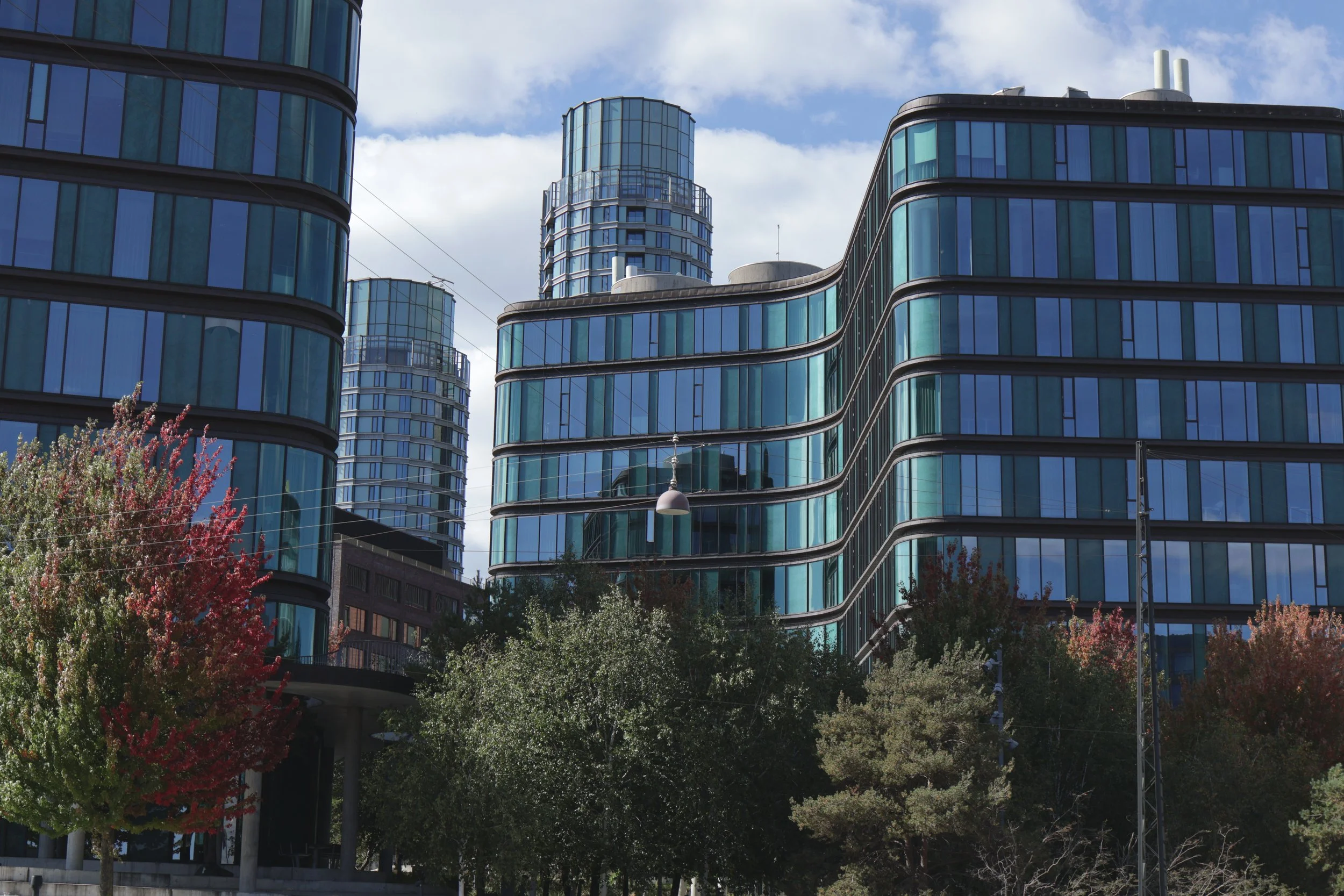 Modern glass buildings with curved facades and round towers, surrounded by trees with colorful fall foliage, under a partly cloudy sky.