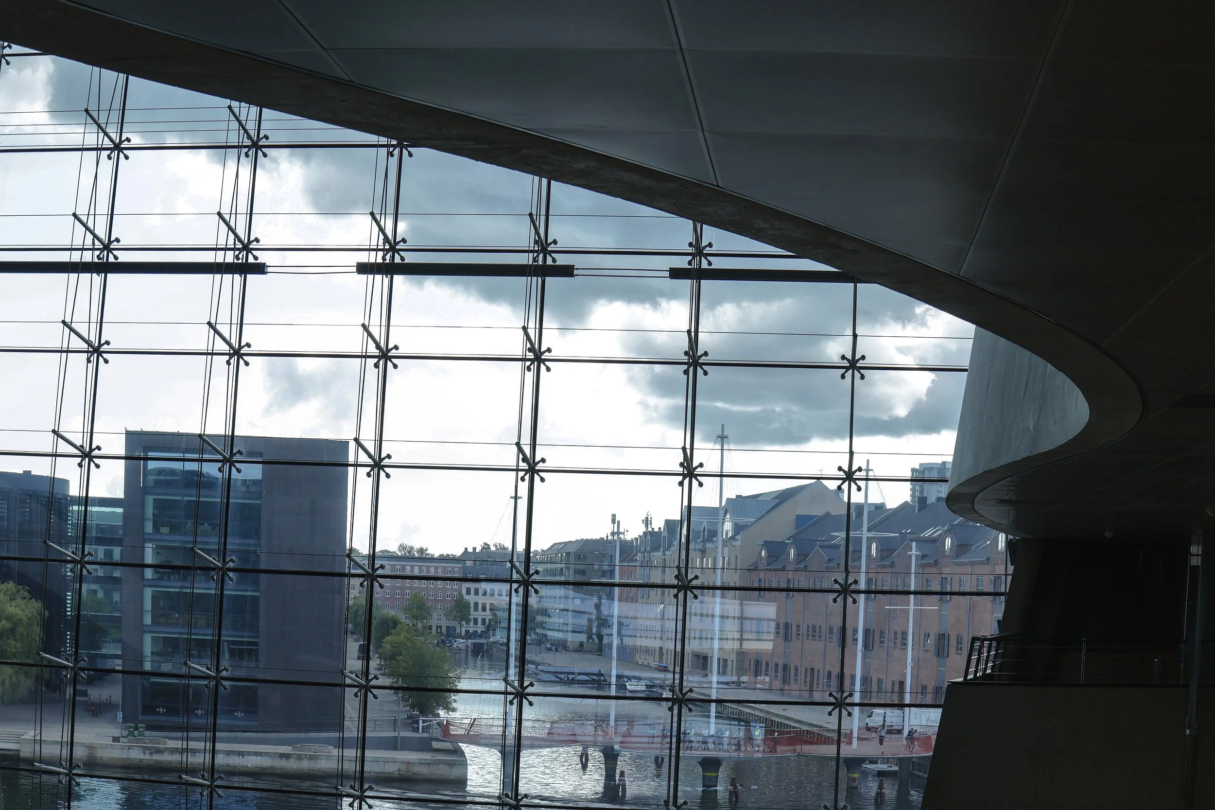 View of modern cityscape through a large glass window with a grid framework, showing buildings, a waterway, and a cloudy sky.