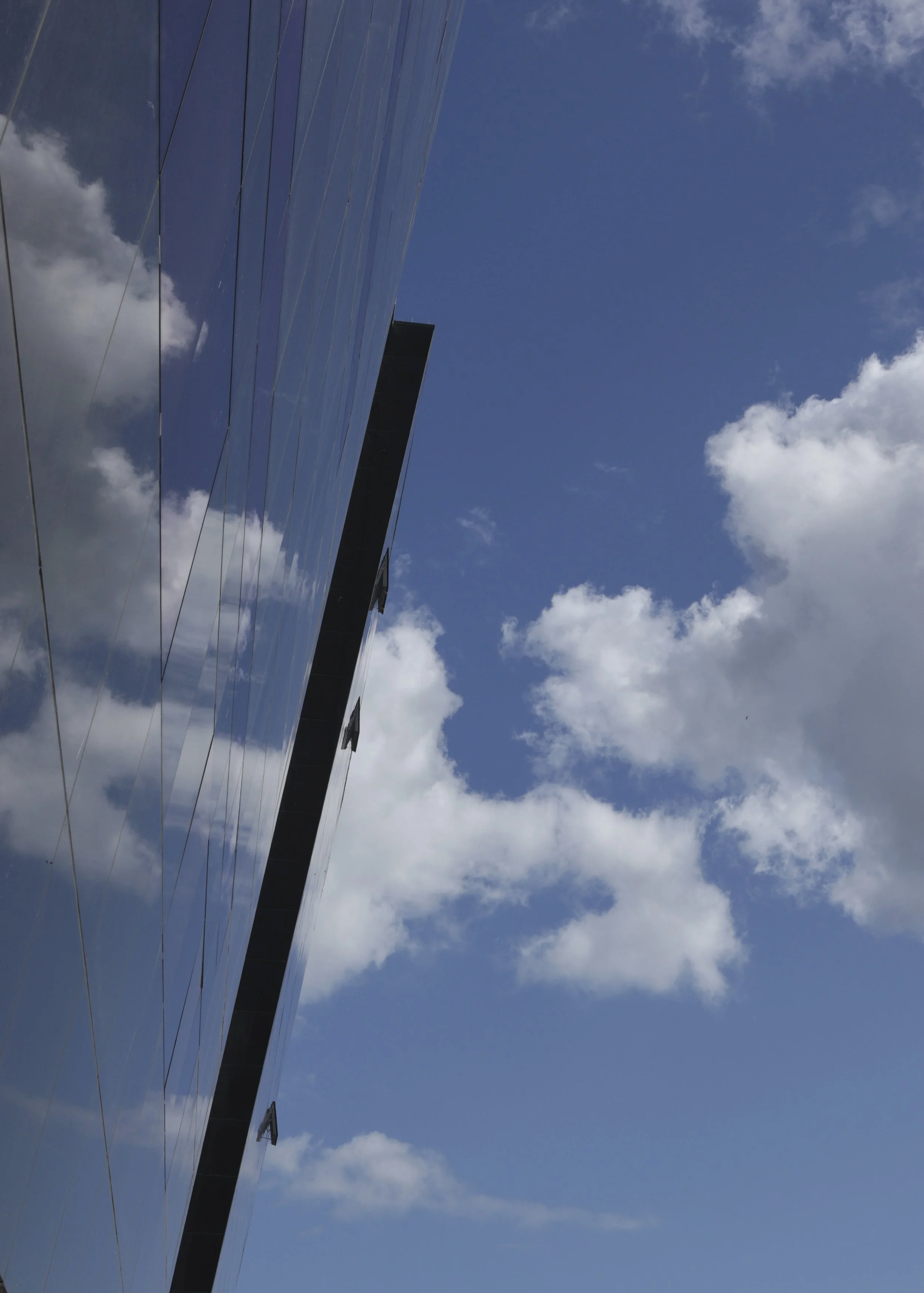A reflective glass building with the sky and clouds reflected on its surface, viewed from below.