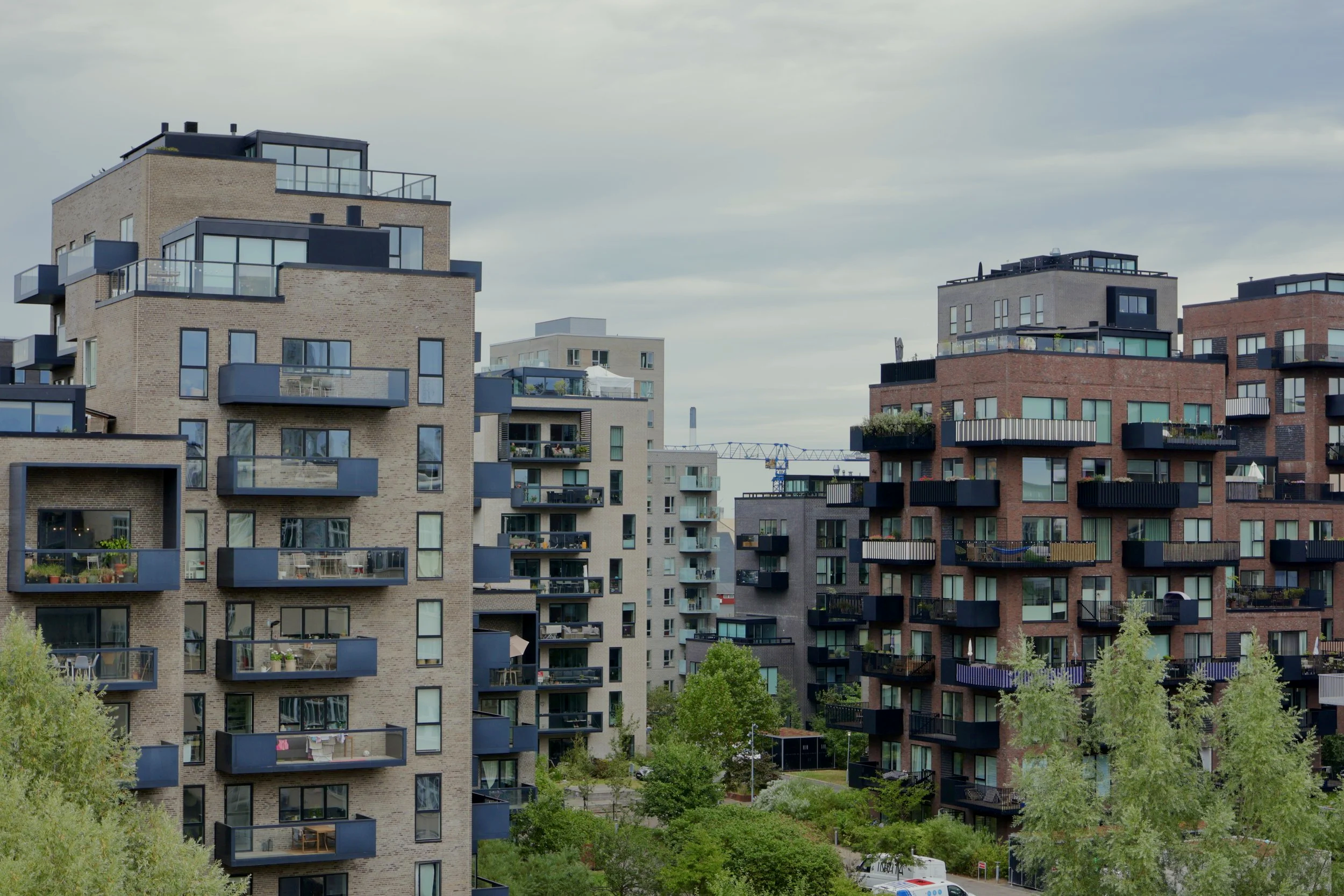Multiple modern apartment buildings with balconies and large windows, surrounded by green trees and a cloudy sky.