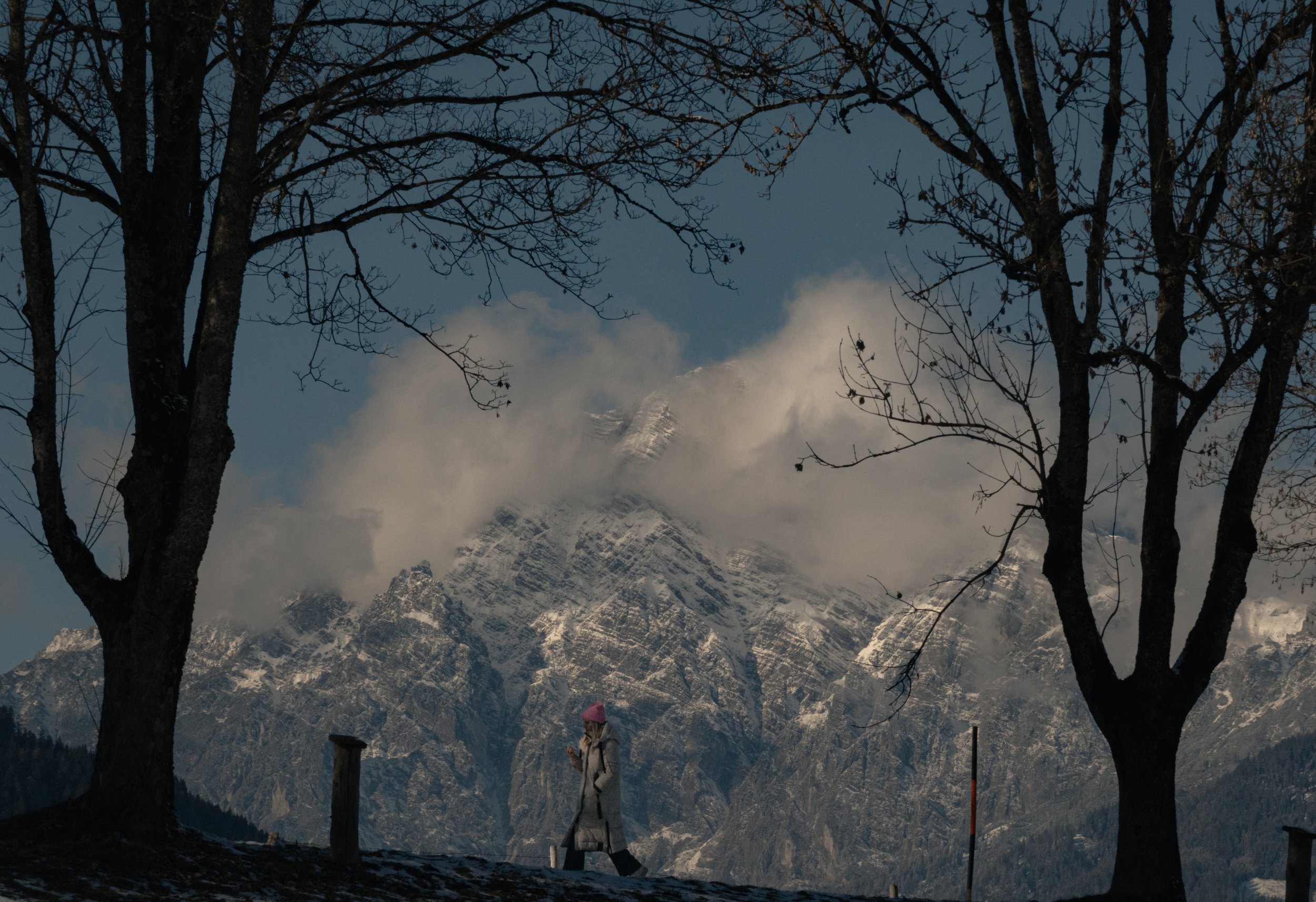 A person walking outdoors near a mountain range with snow, trees in the foreground, and cloudy sky.