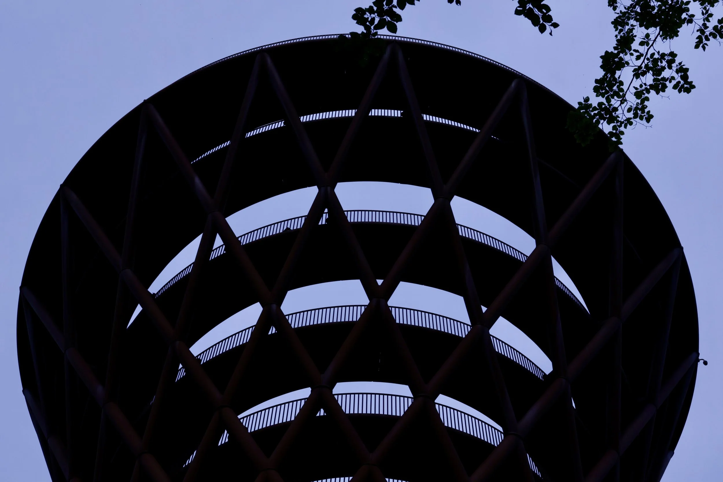 Silhouette of a multi-level observation tower against a blue sky with a few tree branches visible at the top.