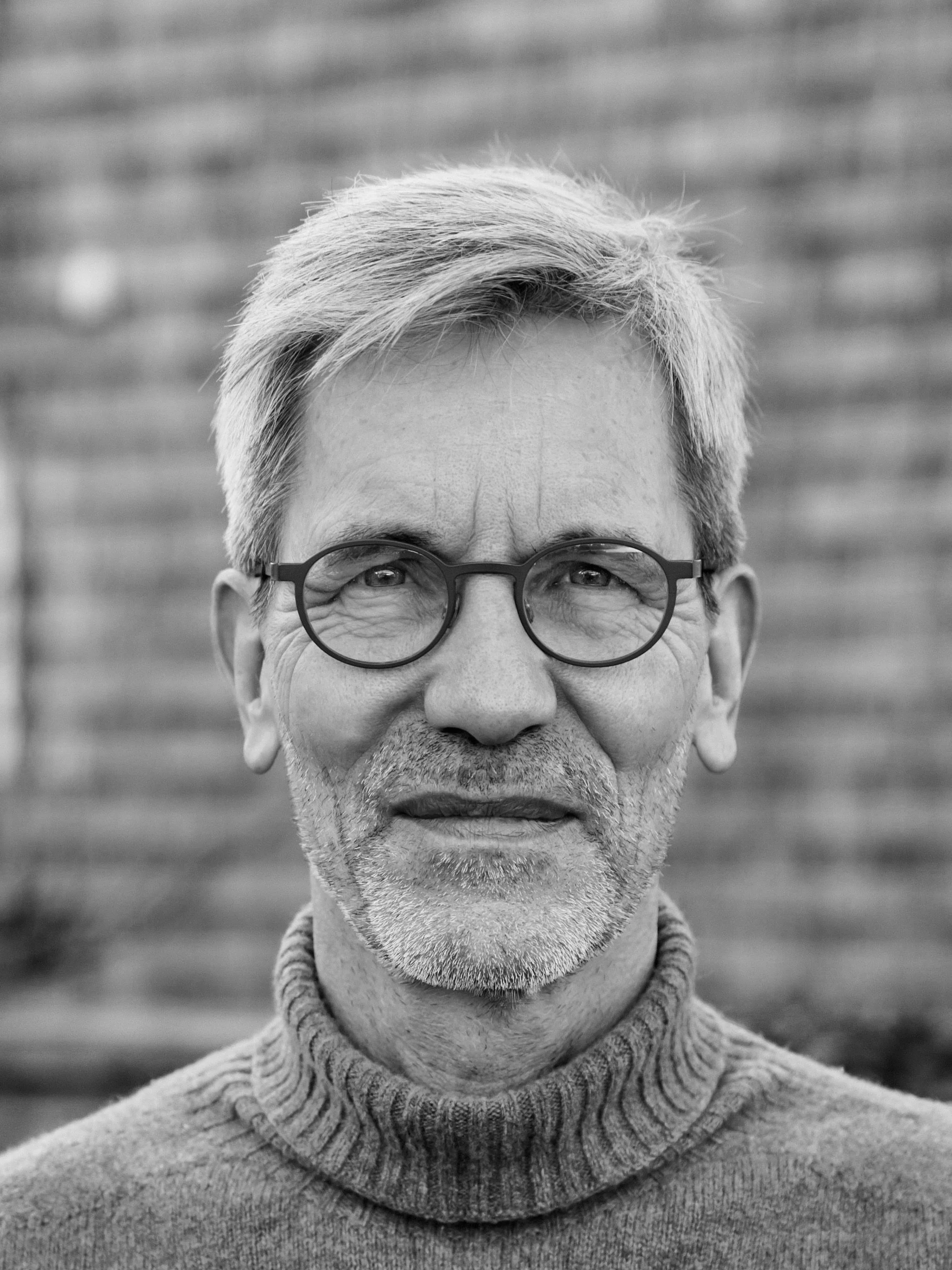 Black and white portrait of a middle-aged man with glasses, gray hair, and a beard, wearing a turtleneck sweater, standing outdoors with a blurred background.