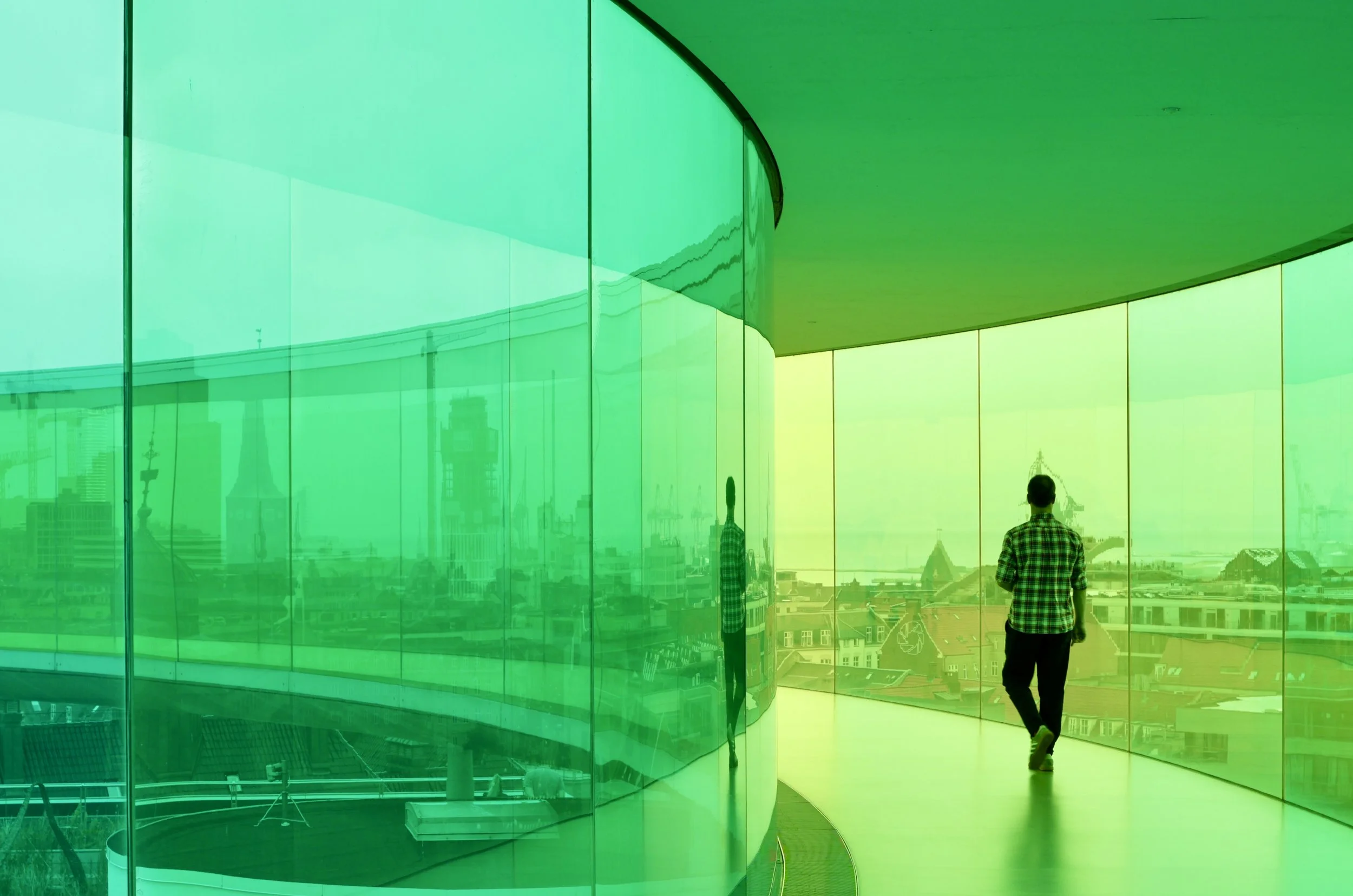 A person in a checkered shirt walking along a curved, glass-walled corridor with a cityscape view in the background.