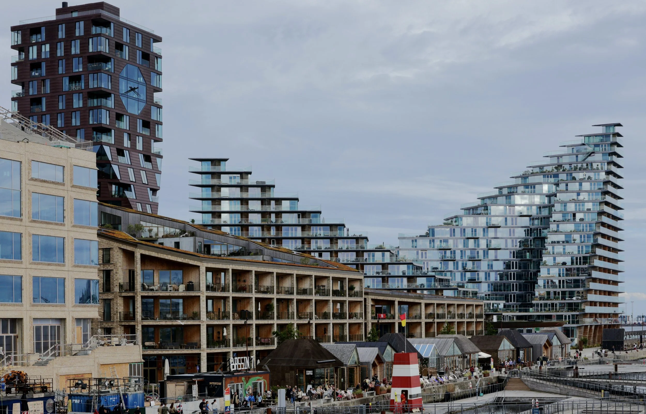 Modern high-rise apartment buildings along the waterfront with balconies, a lighthouse, and small colorful structures, under a cloudy sky.