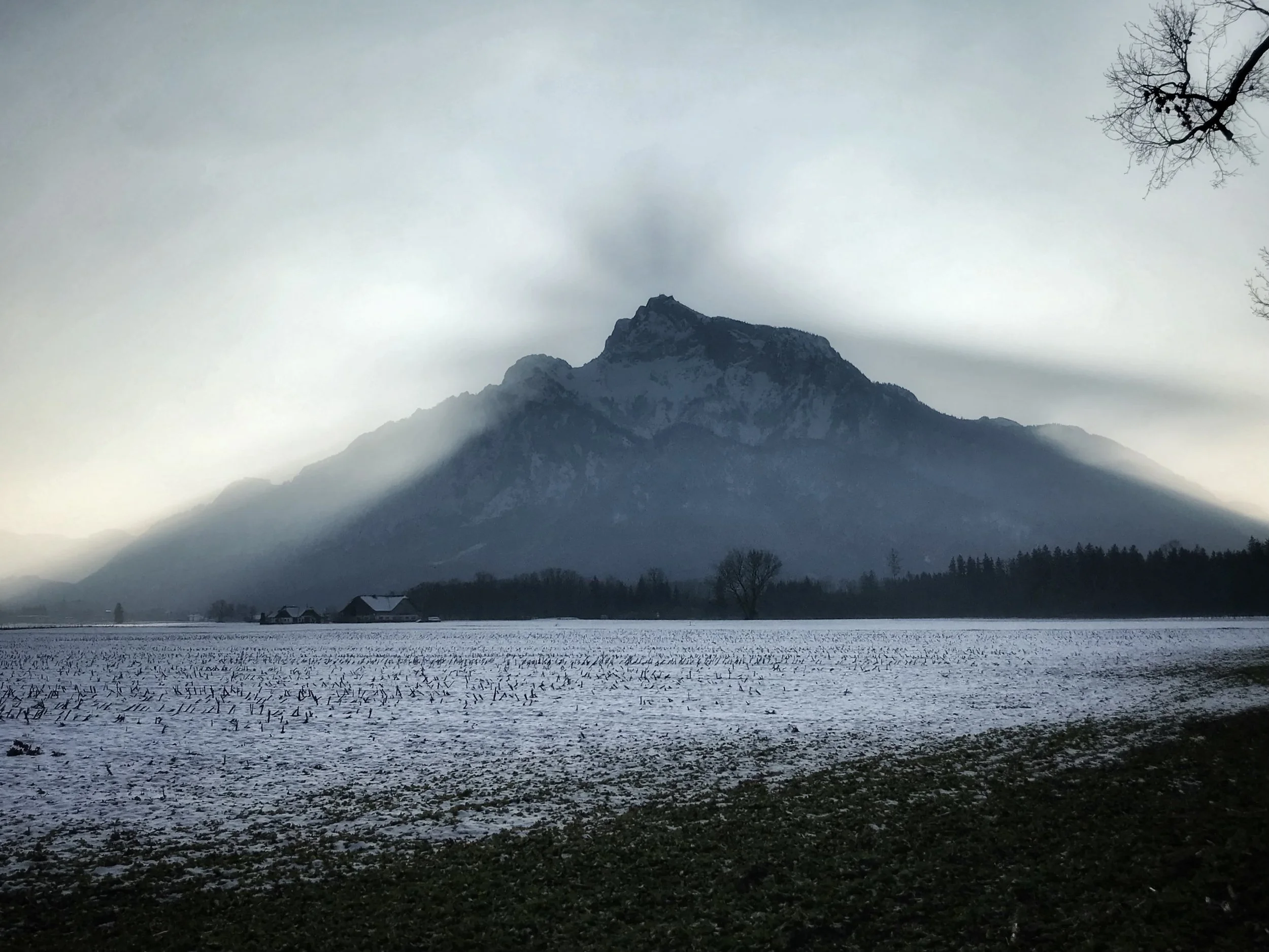Snow-covered field with farm buildings at the base of a tall, rugged mountain partially obscured by clouds