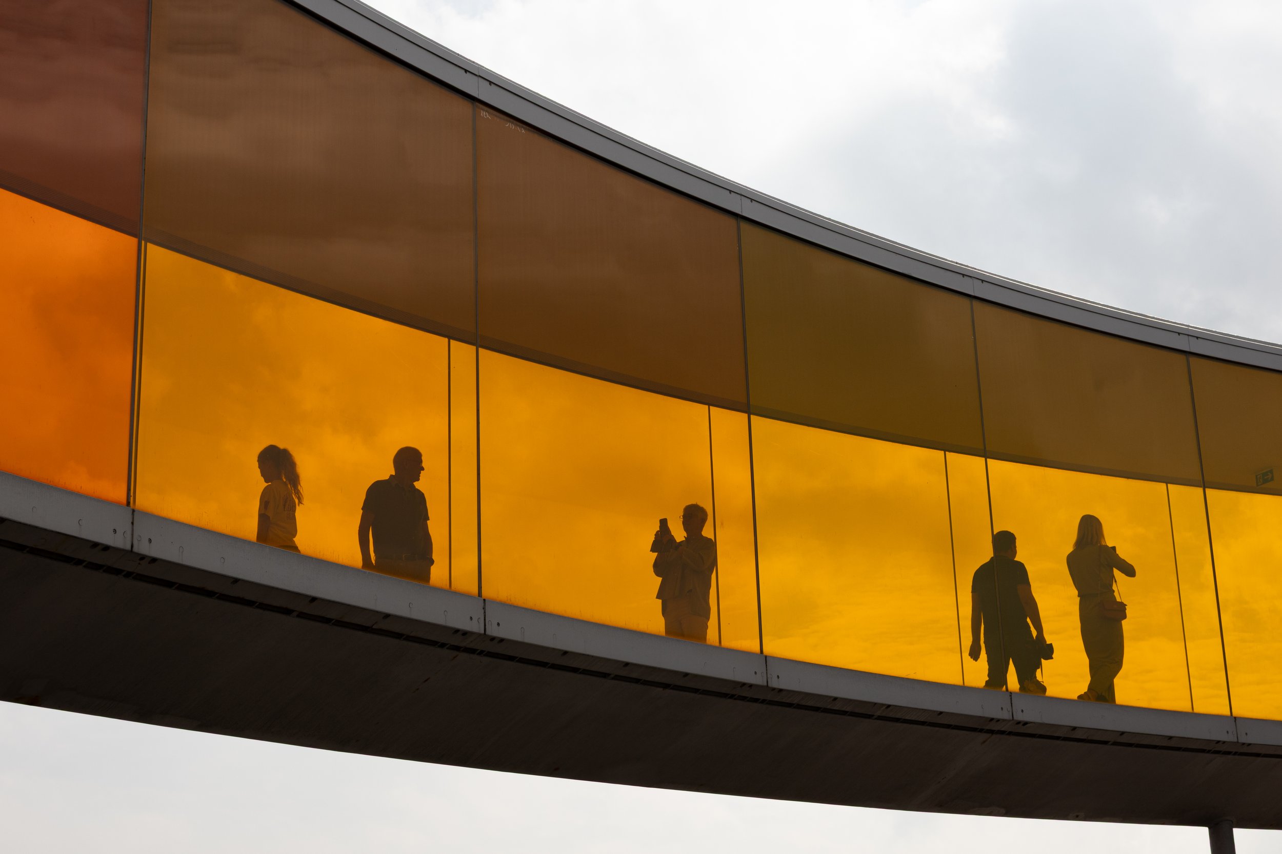 Silhouettes of five people on a skywalk with yellow-tinted glass panels reflecting a cloudy sky.