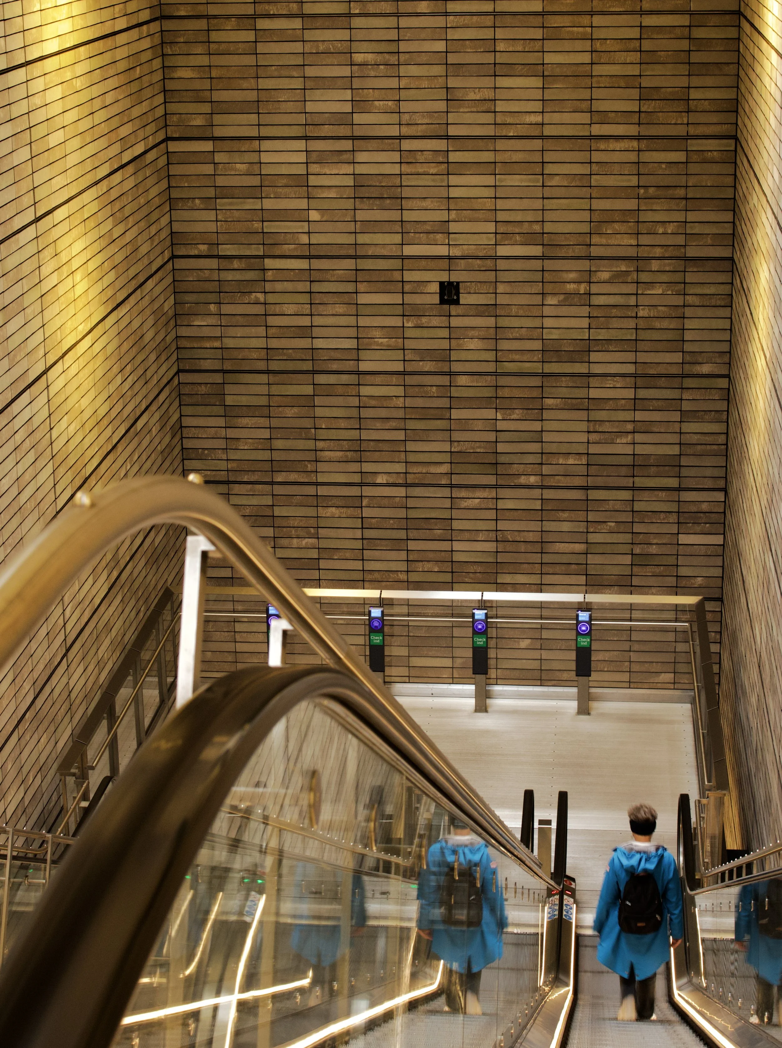 Two people wearing blue jackets and backpacks descending an escalator in a building with brick walls and a tiled ceiling, near a set of electronic check-in or ticket scanners.