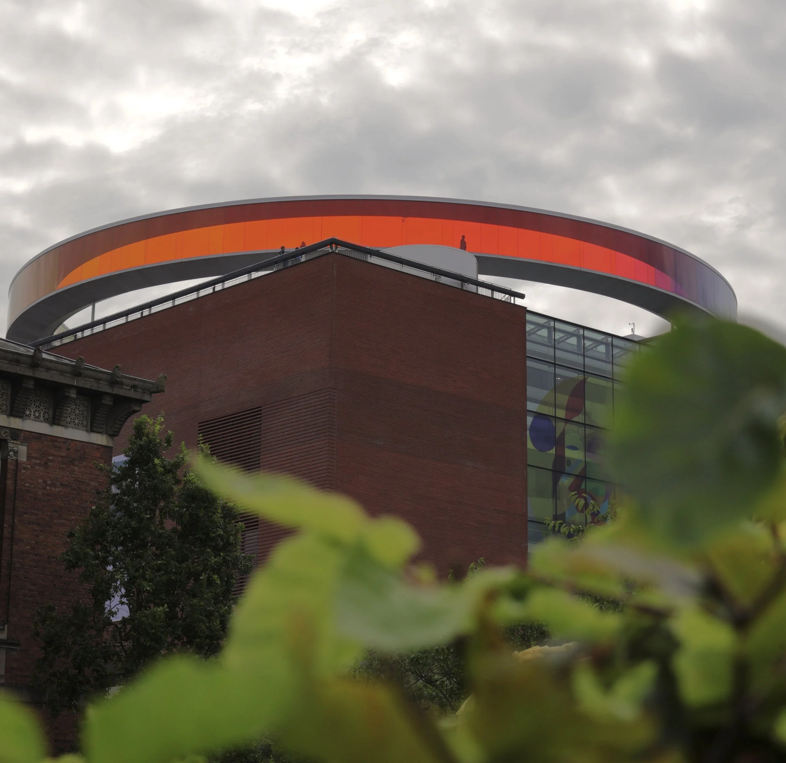 Modern building with a large, circular, orange and purple digital display on top, partially visible in the background, with some greenery in the foreground.